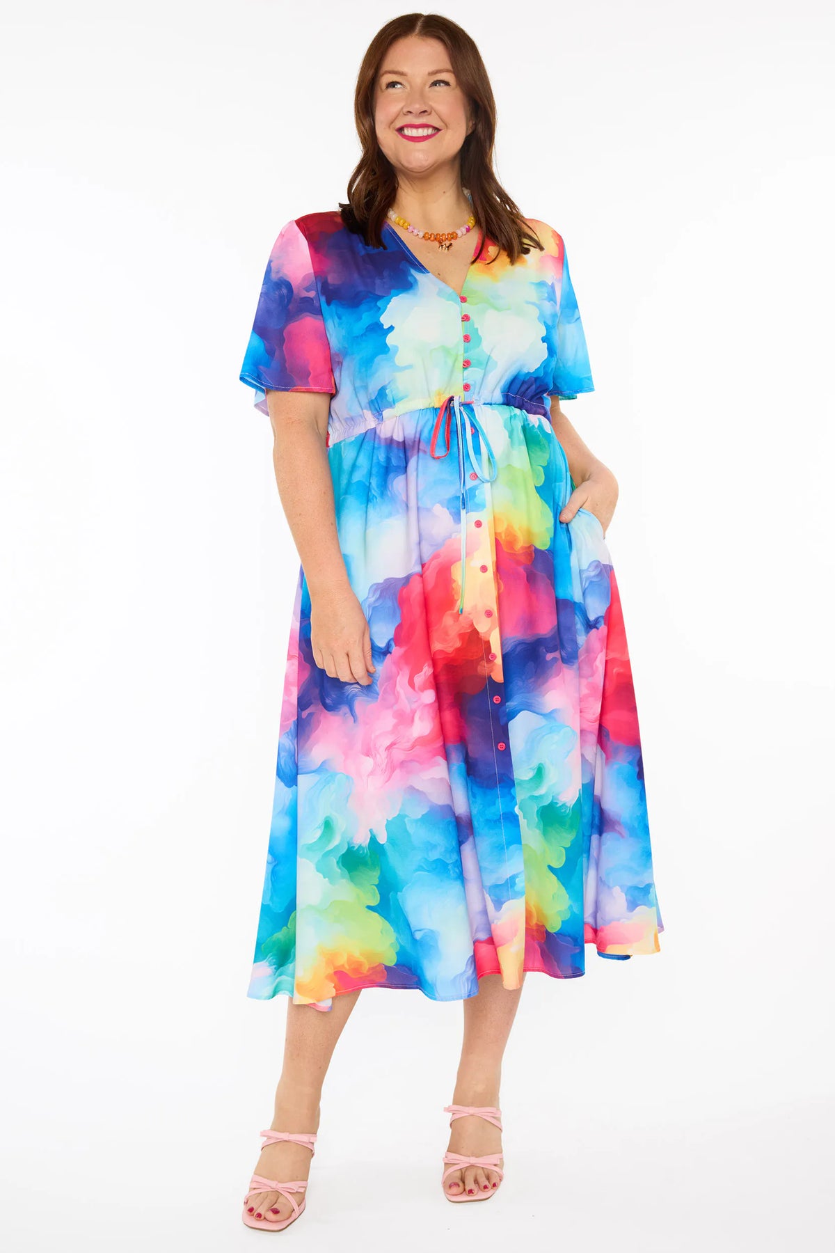 A woman modelling a short-sleeve midi dress with a vibrant watercolour tie-dye pattern in shades of blue, pink, yellow, green, and purple, set against a plain white studio background.