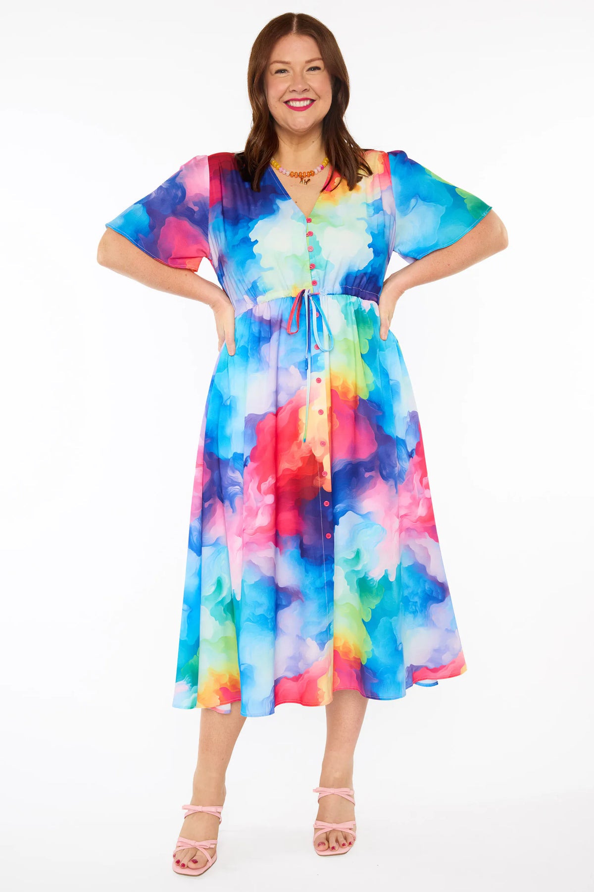 A woman modelling a short-sleeve midi dress with a vibrant watercolour tie-dye pattern in shades of blue, pink, yellow, green, and purple, set against a plain white studio background.