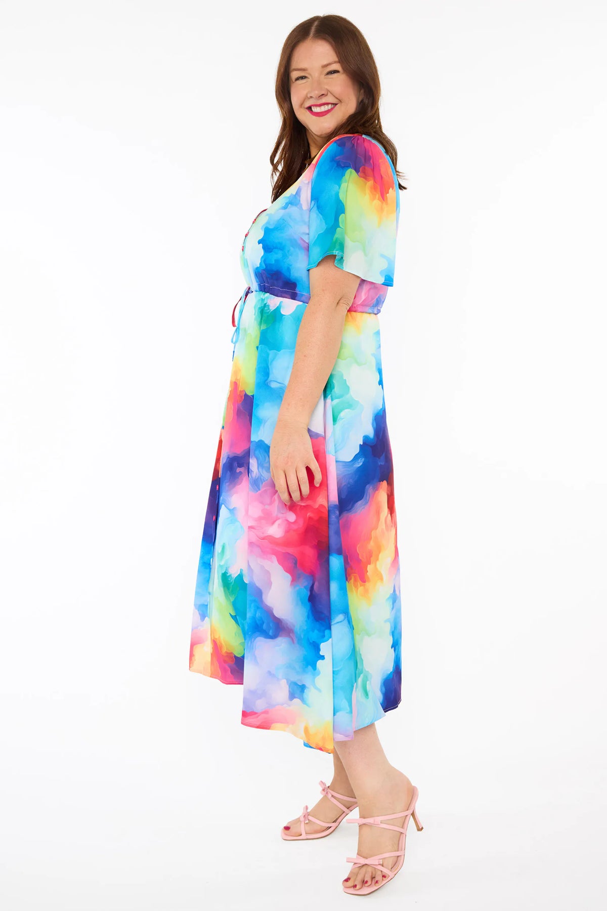 A woman modelling a short-sleeve midi dress with a vibrant watercolour tie-dye pattern in shades of blue, pink, yellow, green, and purple, set against a plain white studio background.