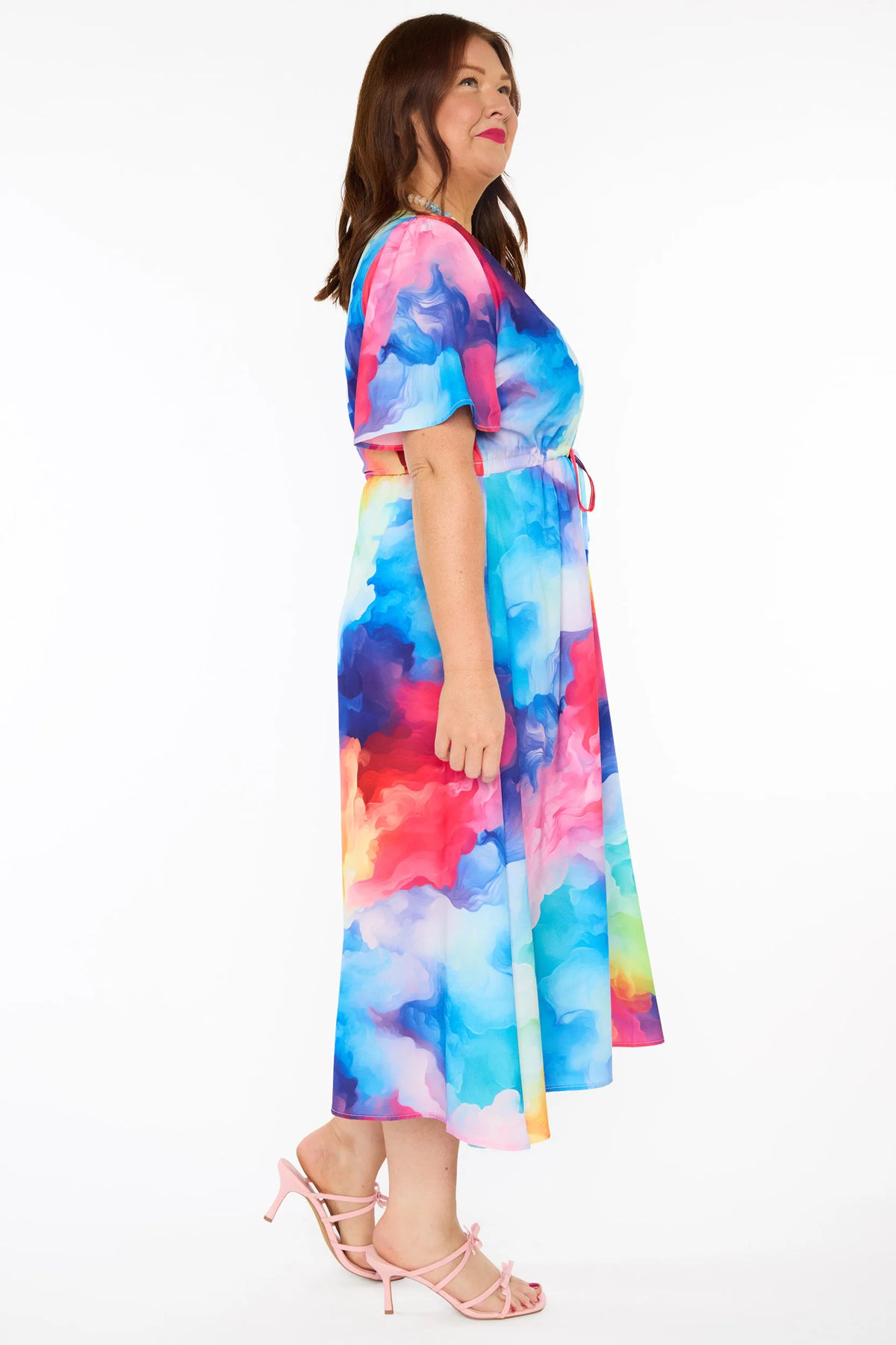 A woman modelling a short-sleeve midi dress with a vibrant watercolour tie-dye pattern in shades of blue, pink, yellow, green, and purple, set against a plain white studio background.