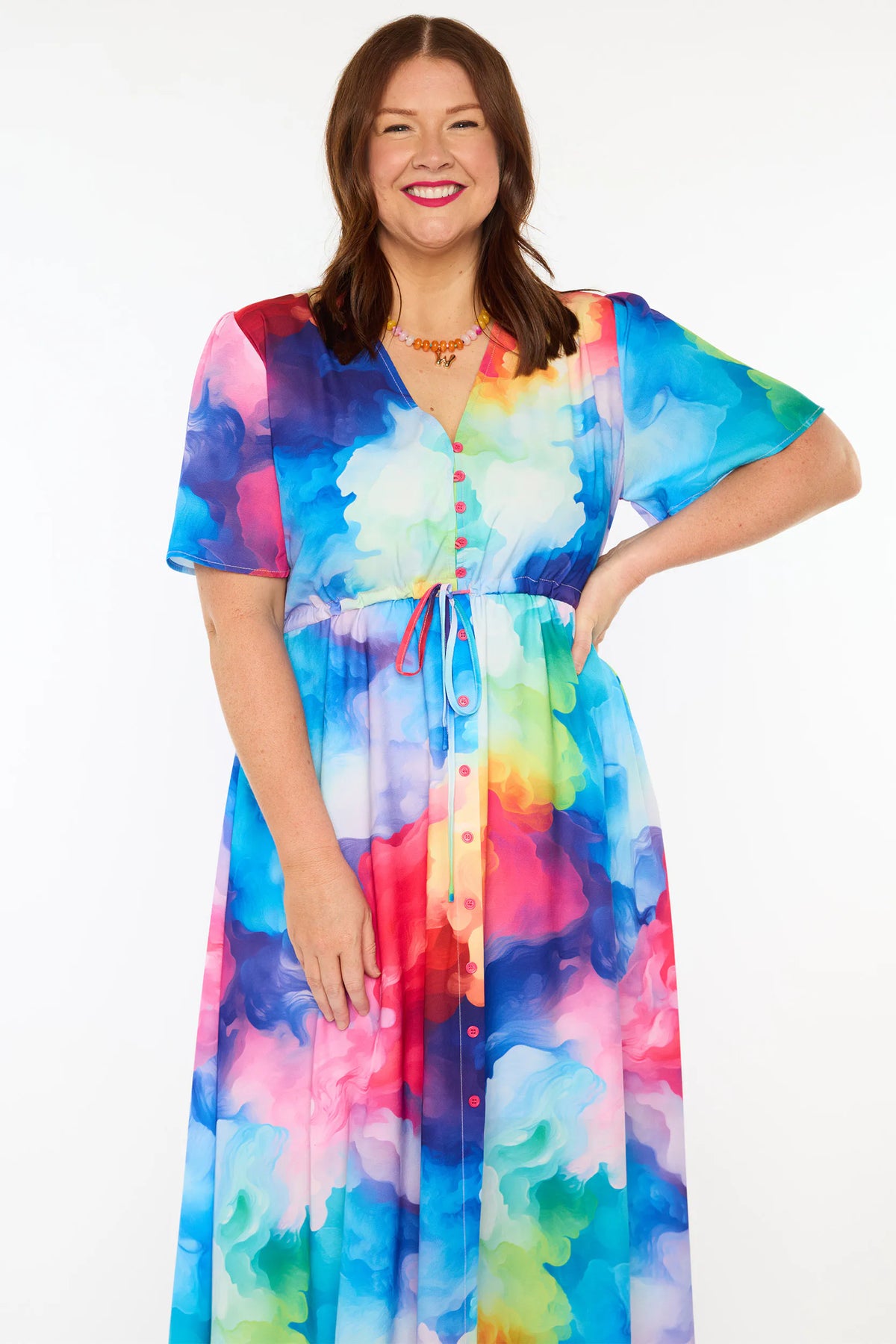 A woman modelling a short-sleeve midi dress with a vibrant watercolour tie-dye pattern in shades of blue, pink, yellow, green, and purple, set against a plain white studio background.