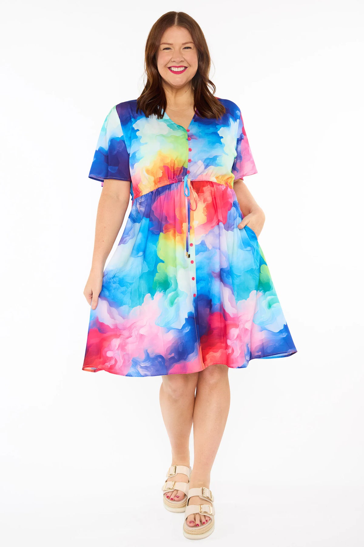 A woman modelling a short-sleeve V neckline button-front dress with a vibrant rainbow cloud pattern, standing against a plain white studio background.