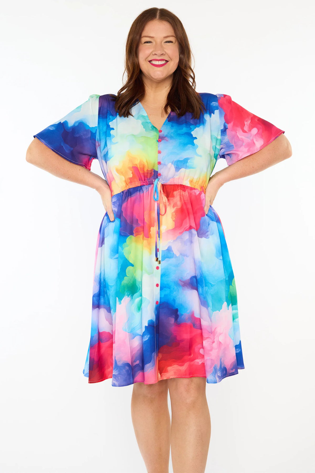A woman modelling a short-sleeve V neckline button-front dress with a vibrant rainbow cloud pattern, standing against a plain white studio background.