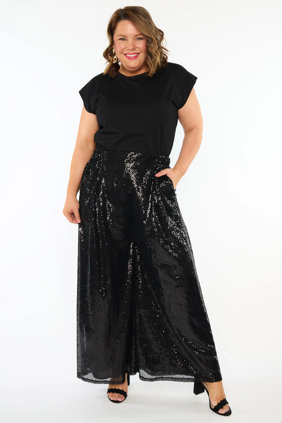 A woman modelling a black short sleeves top and a black sequin wide leg pant, standing against a plain white studio background with black heels.