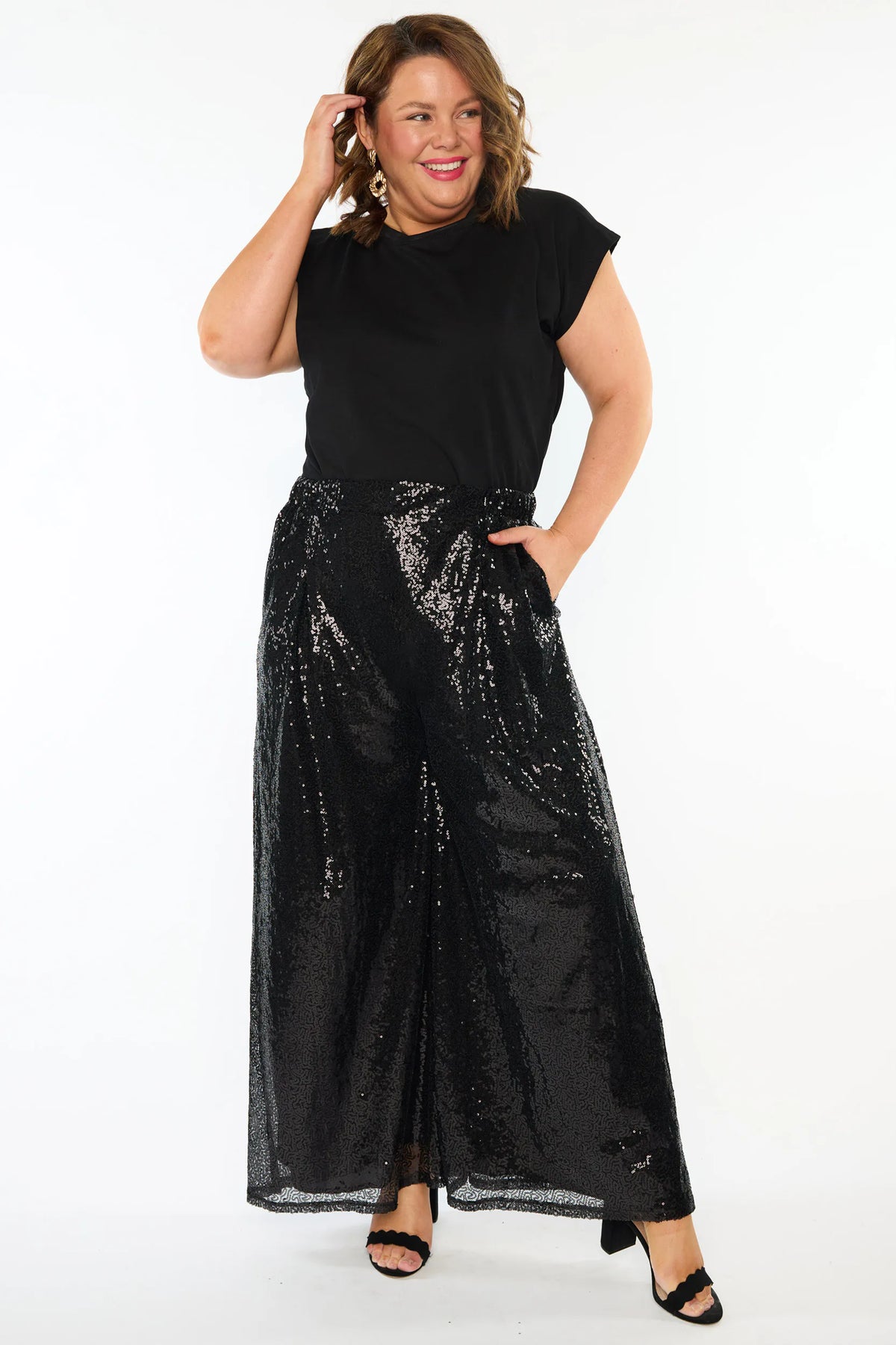 A woman modelling a black short sleeves top and a black sequin wide leg pant, standing against a plain white studio background with black heels.