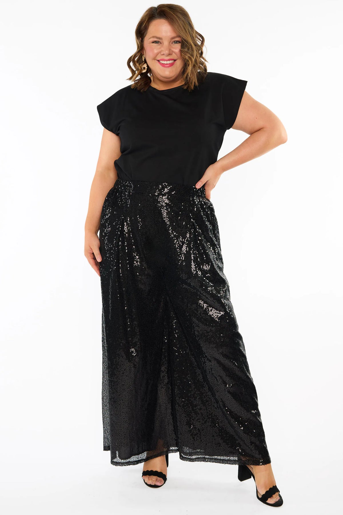A woman modelling a black short sleeves top and a black sequin wide leg pant, standing against a plain white studio background with black heels.