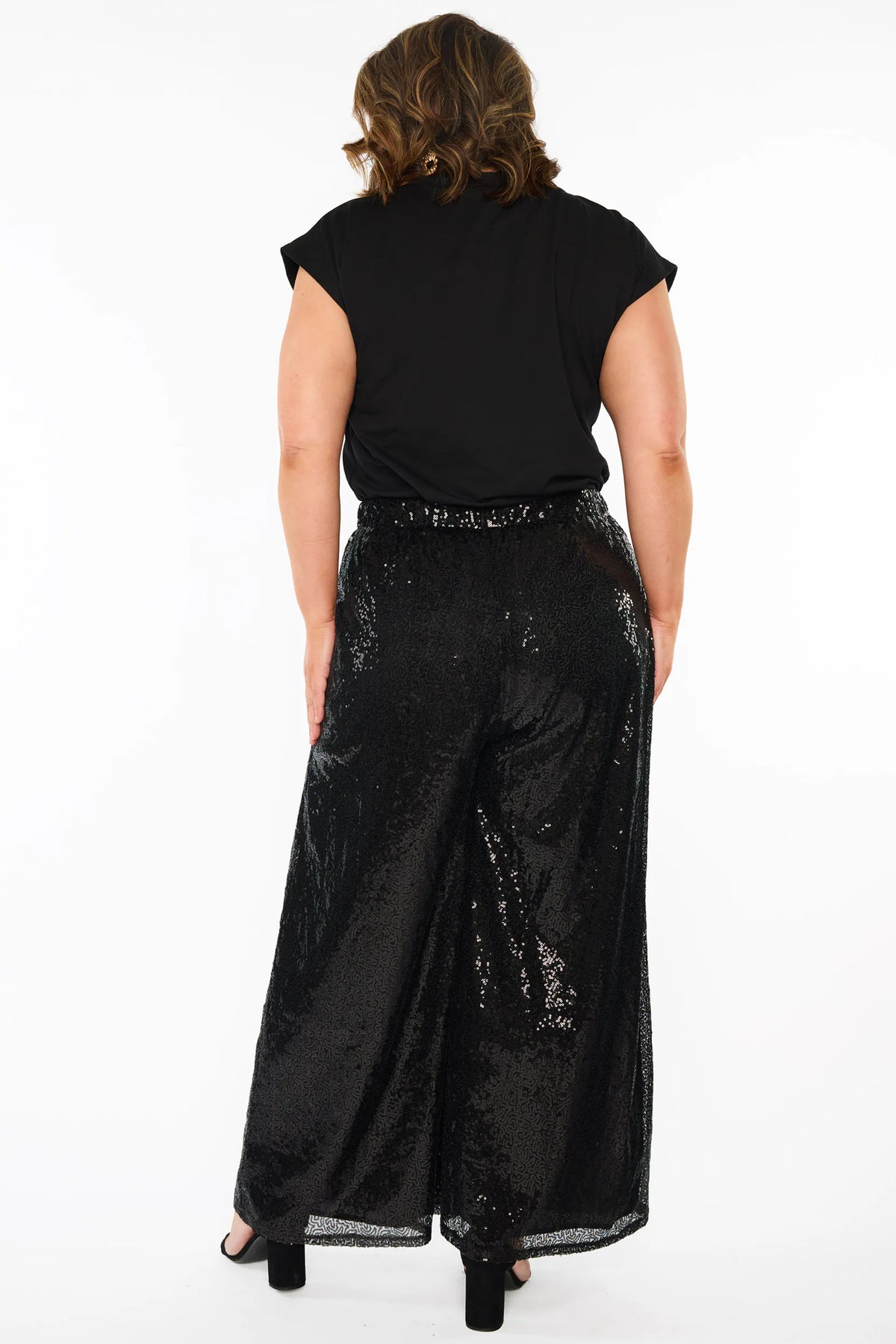 A woman modelling a black short sleeves top and a black sequin wide leg pant, standing against a plain white studio background with black heels.