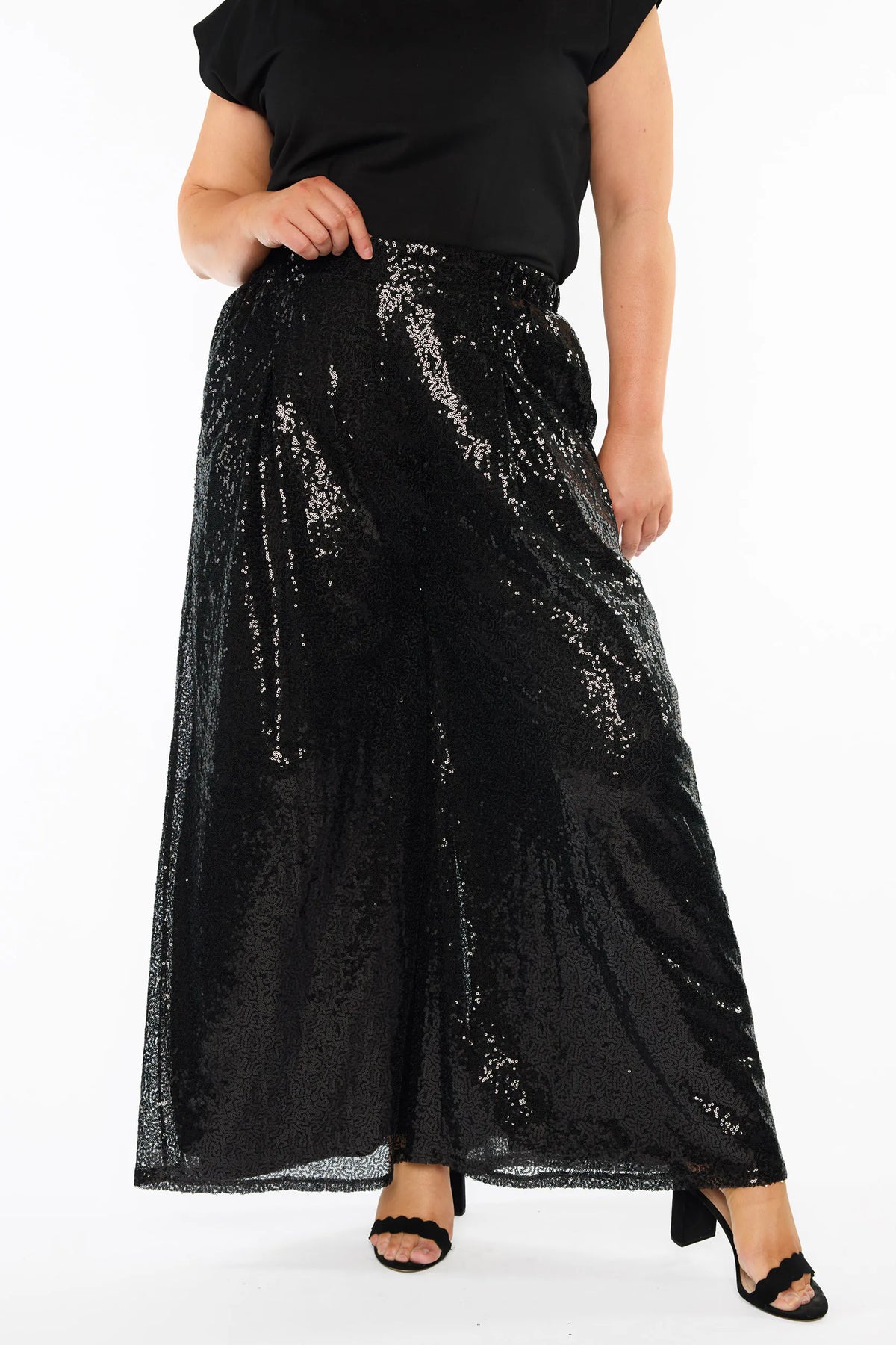 Close up of a woman modelling a black short sleeves top and a black sequin wide leg pant, standing against a plain white studio background with black heels.
