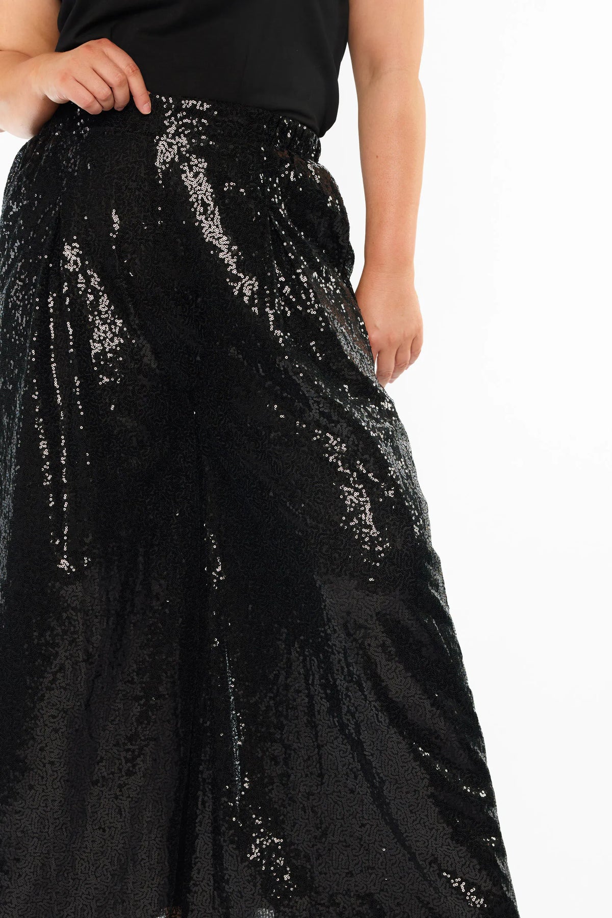 A close up of a woman modelling a black short sleeves top and a black sequin wide leg pant, standing against a plain white studio background with black heels.
