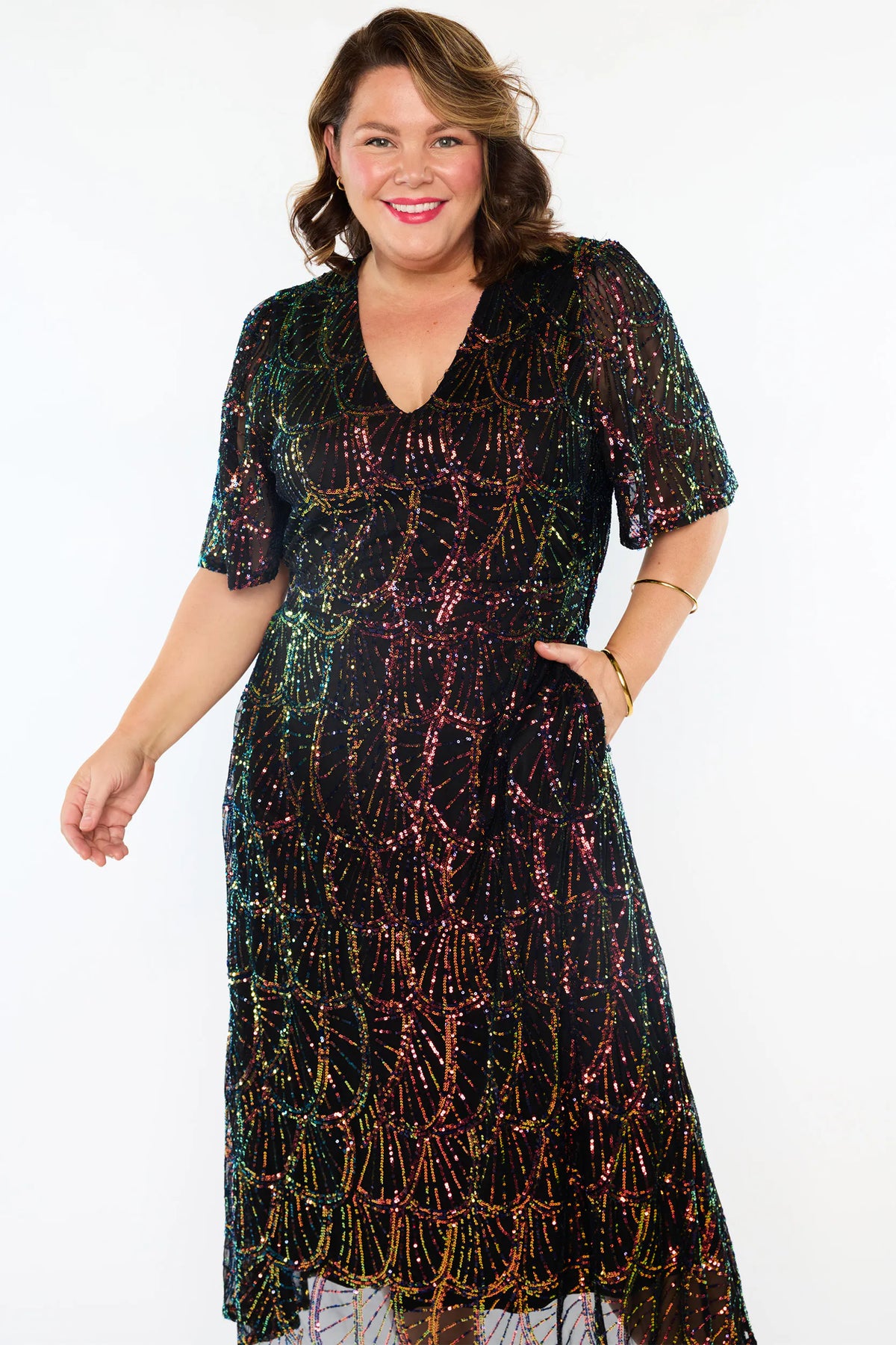 A woman modeling a short-sleeve V-neck midi dress covered in iridescent sequins that shift between green, pink, purple, and gold on a plain white background.