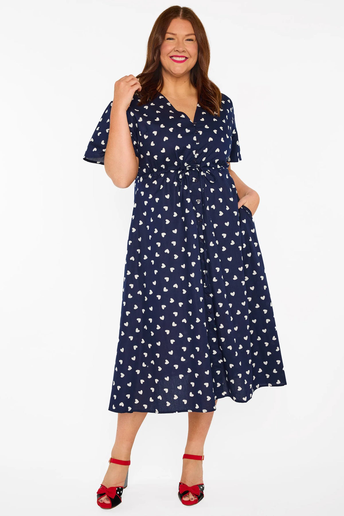 A woman modelling a navy blue midi dress with a white heart pattern, with V-neckline, short sleeves and an adjustable tie waist, standing against a plain white studio background.
