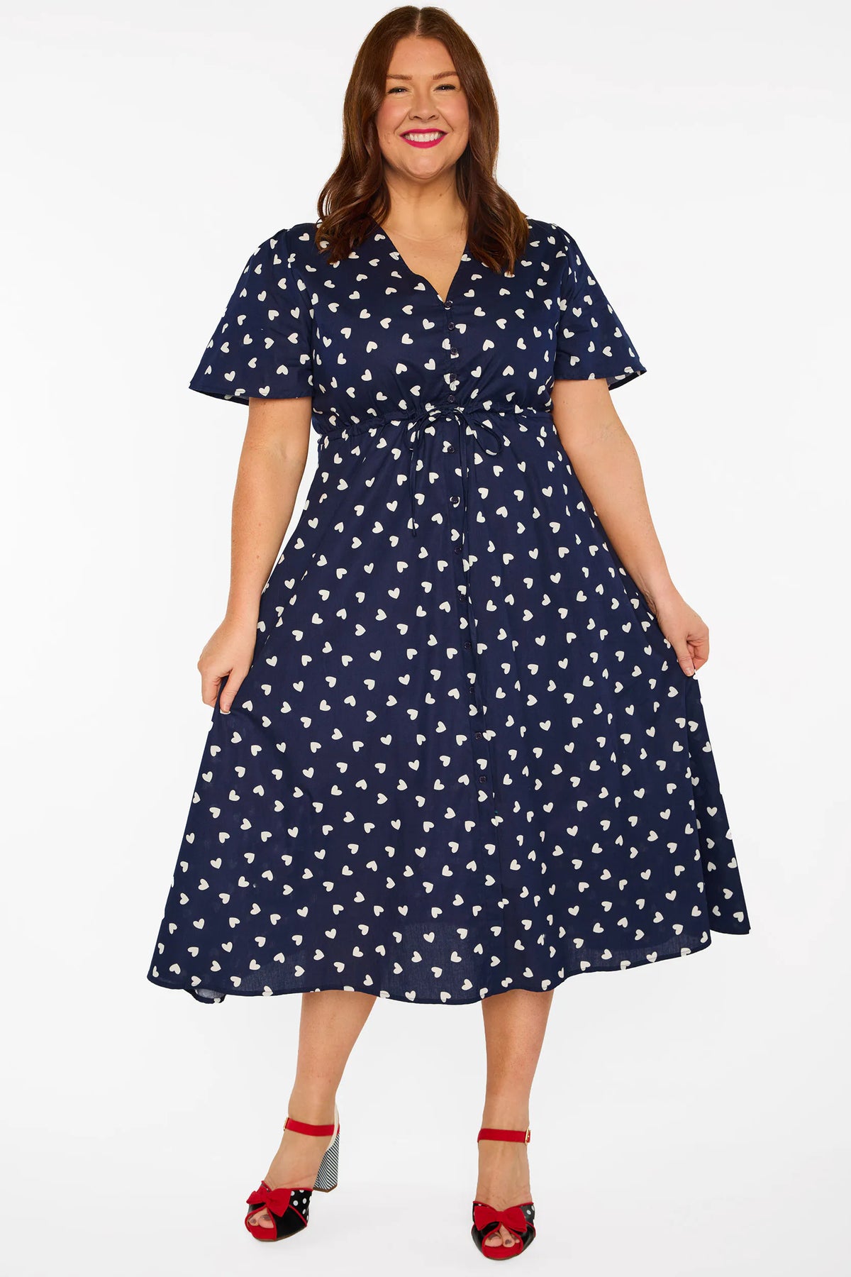 A woman modelling a navy blue midi dress with a white heart pattern, with V-neckline, short sleeves and an adjustable tie waist, standing against a plain white studio background.