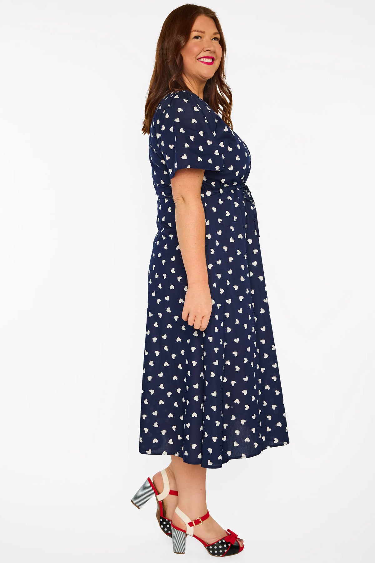 A woman modelling a navy blue midi dress with a white heart pattern, with V-neckline, short sleeves and an adjustable tie waist, standing against a plain white studio background.