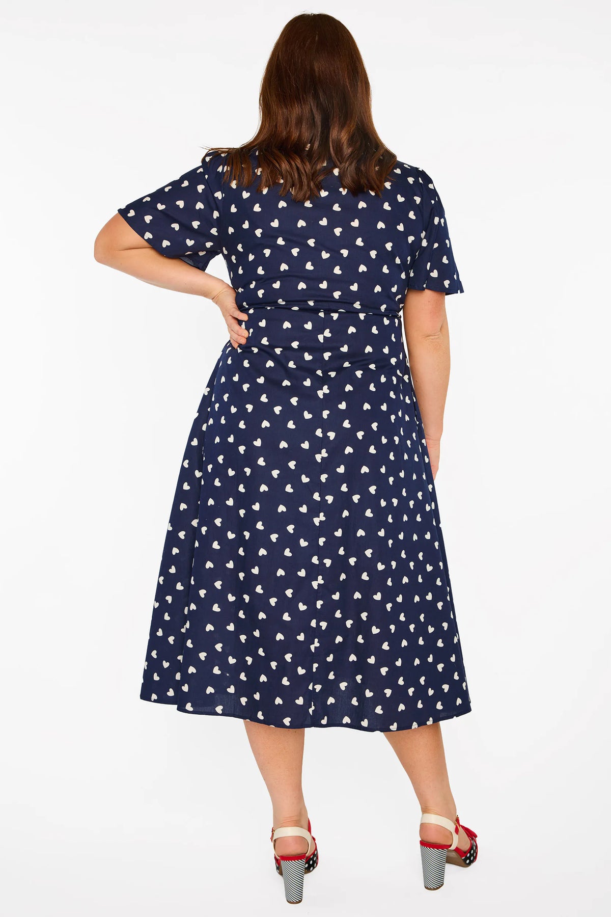 A woman modelling a navy blue midi dress with a white heart pattern, with V-neckline, short sleeves and an adjustable tie waist, standing against a plain white studio background.
