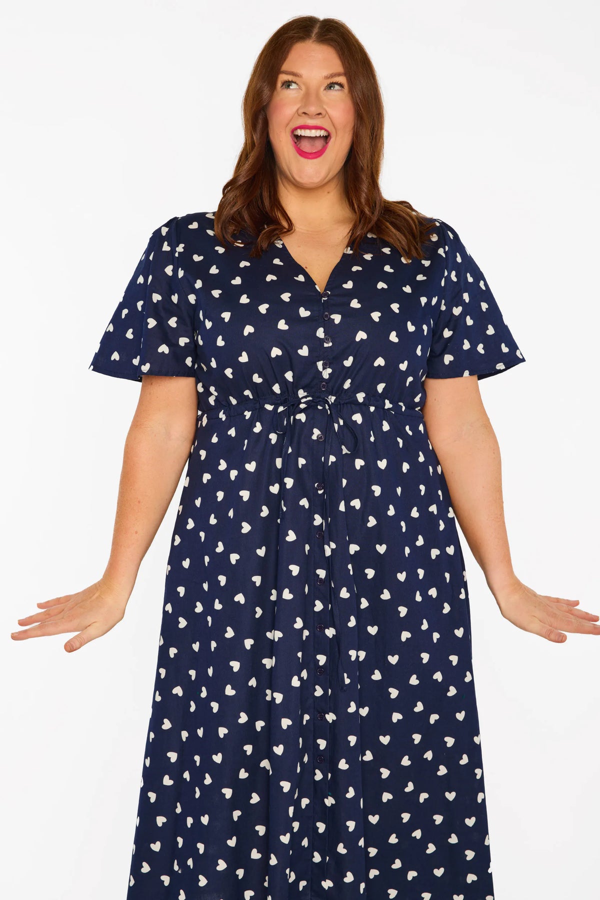 A woman modelling a navy blue midi dress with a white heart pattern, with V-neckline, short sleeves and an adjustable tie waist, standing against a plain white studio background.