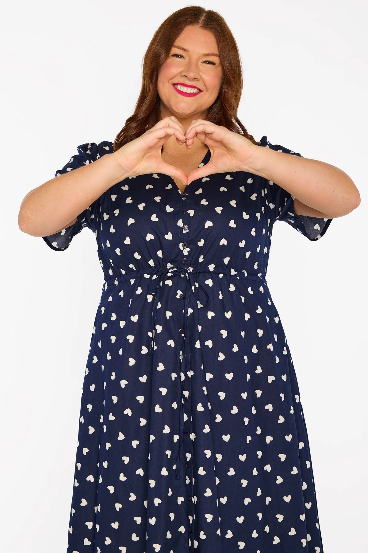 A woman modelling a navy blue midi dress with a white heart pattern, with V-neckline, short sleeves and an adjustable tie waist, standing against a plain white studio background.