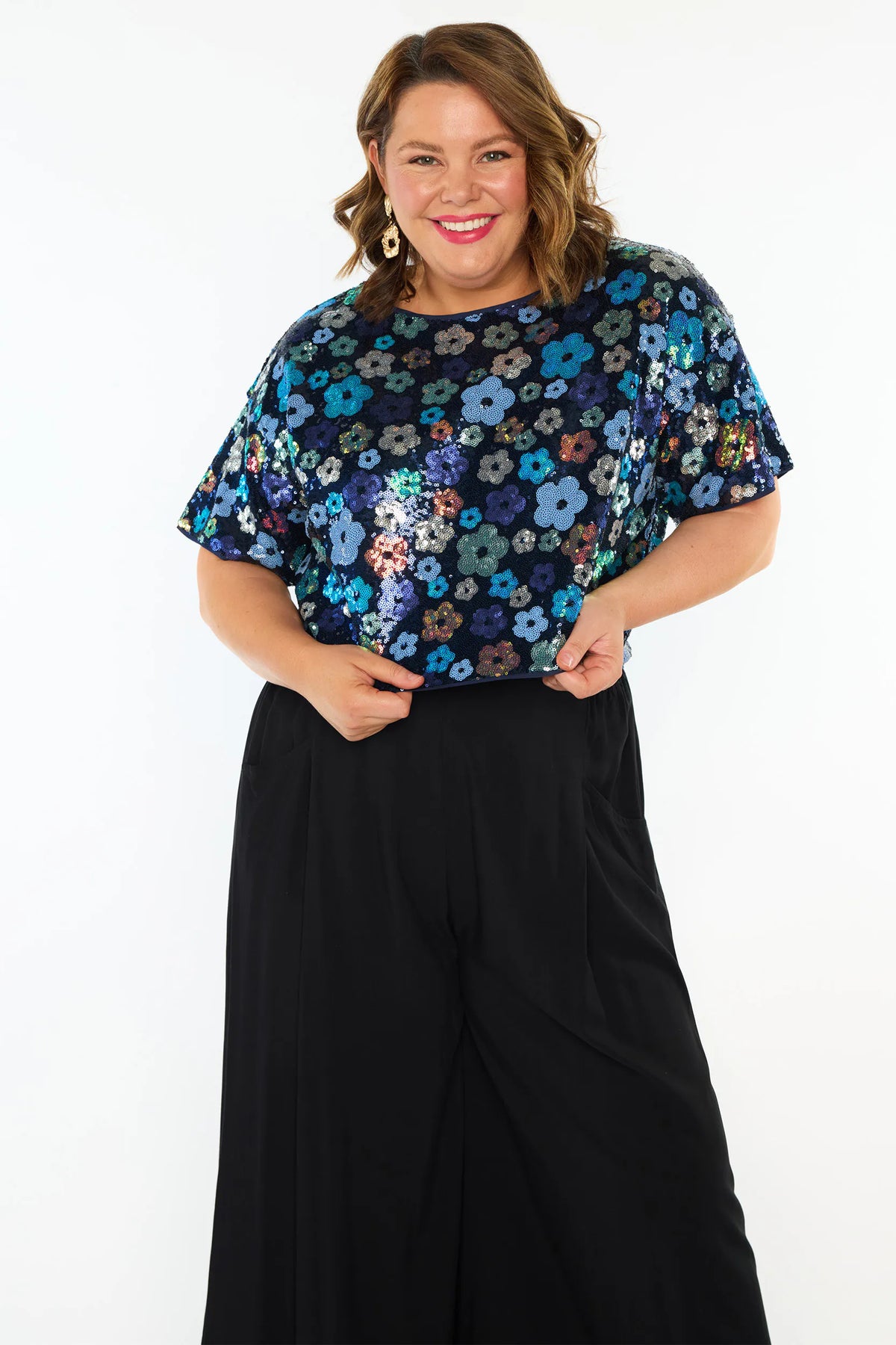 A woman modelling a short-sleeve top with a mix of blue tonal coloured sequin flowers on a black sequin background, paired with black wide-leg pants and black heeled sandals, against a plain white studio background.