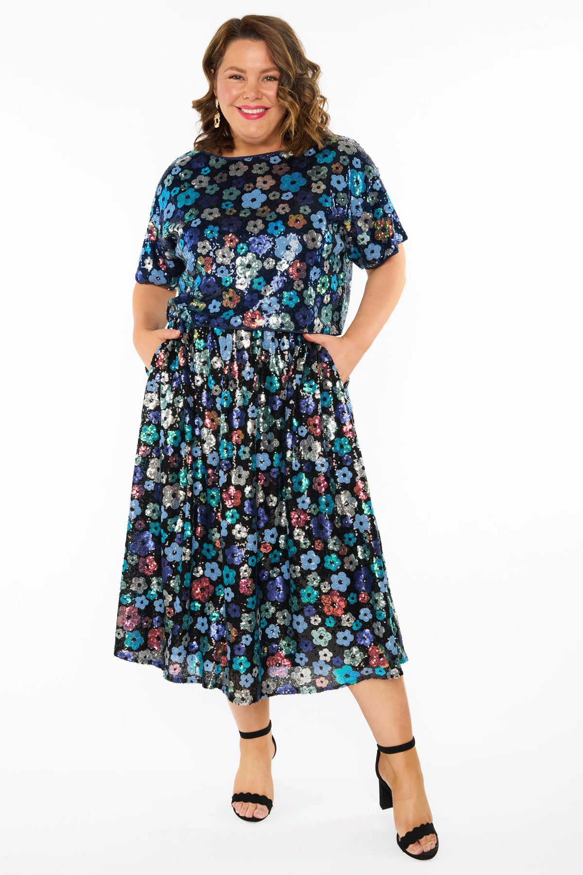 A woman modelling a sequins short-sleeve top with a mix of blue tonal coloured flowers on a black background, paired with a matching knee length skirt and black heels, against a plain white studio background.