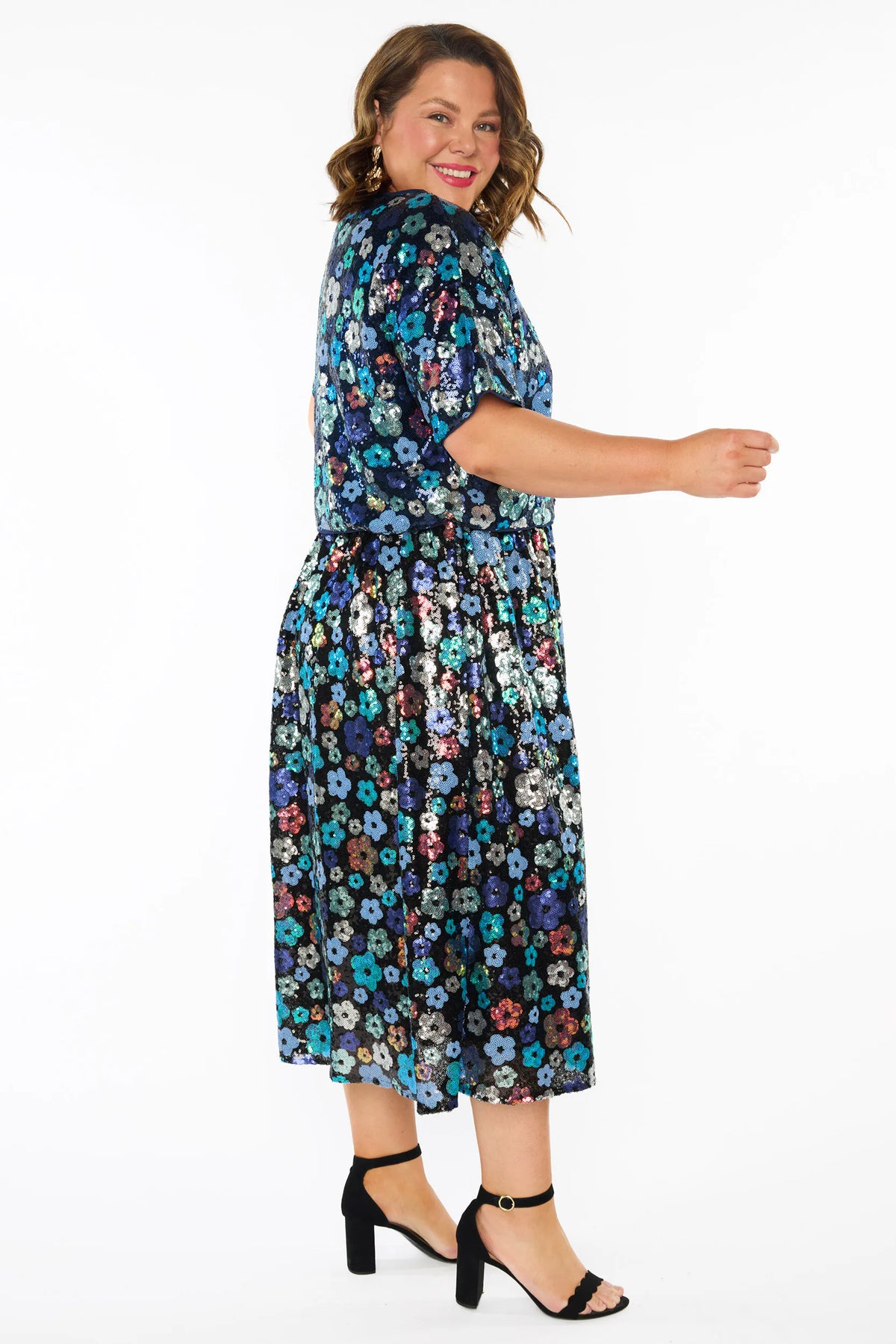 A woman modelling a sequins short-sleeve top with a mix of blue tonal coloured flowers on a black background, paired with a matching knee length skirt and black heels, against a plain white studio background.