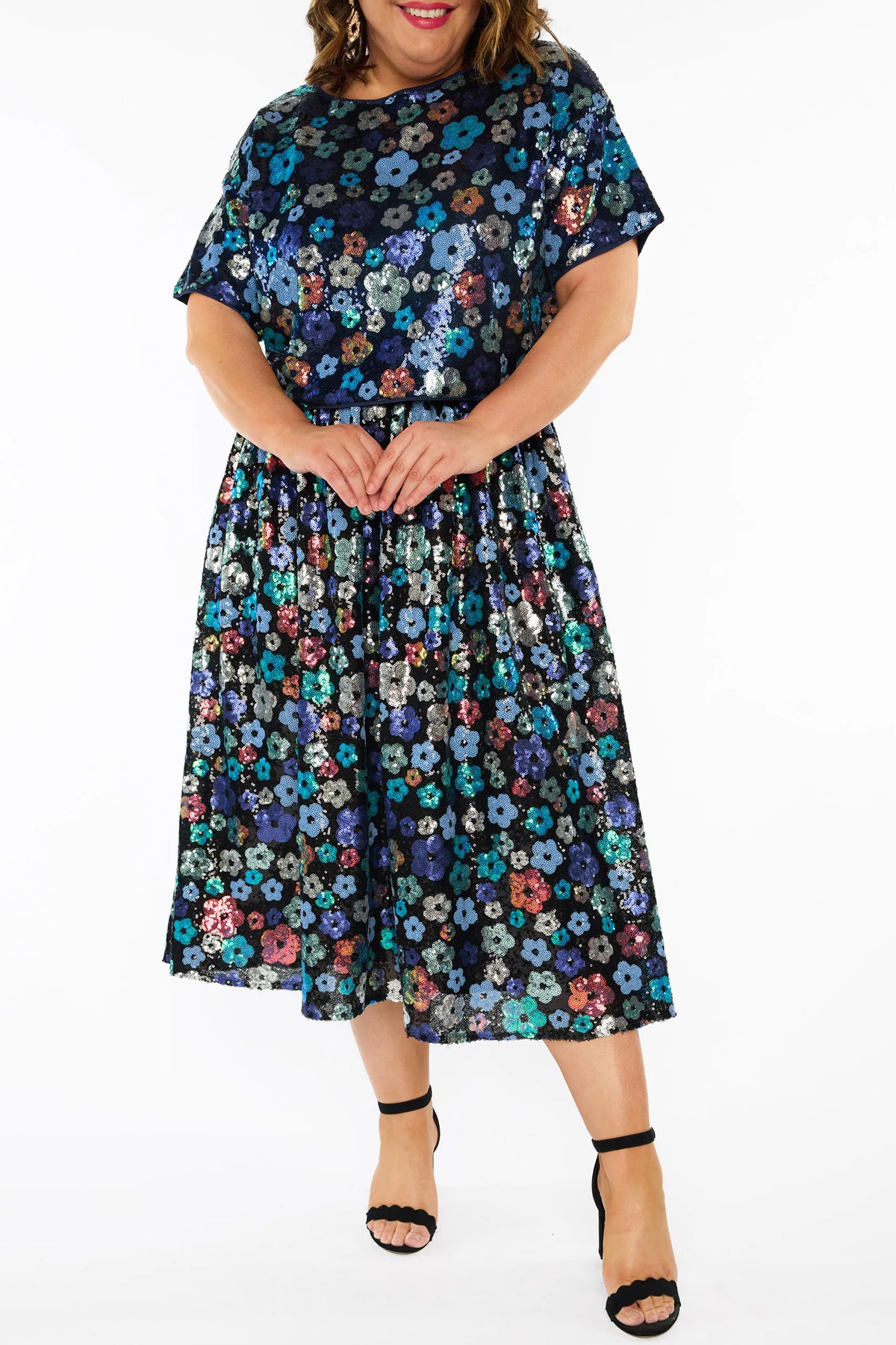 A woman modelling a sequins short-sleeve top with a mix of blue tonal coloured flowers on a black background, paired with a matching knee length skirt and black heels, against a plain white studio background.