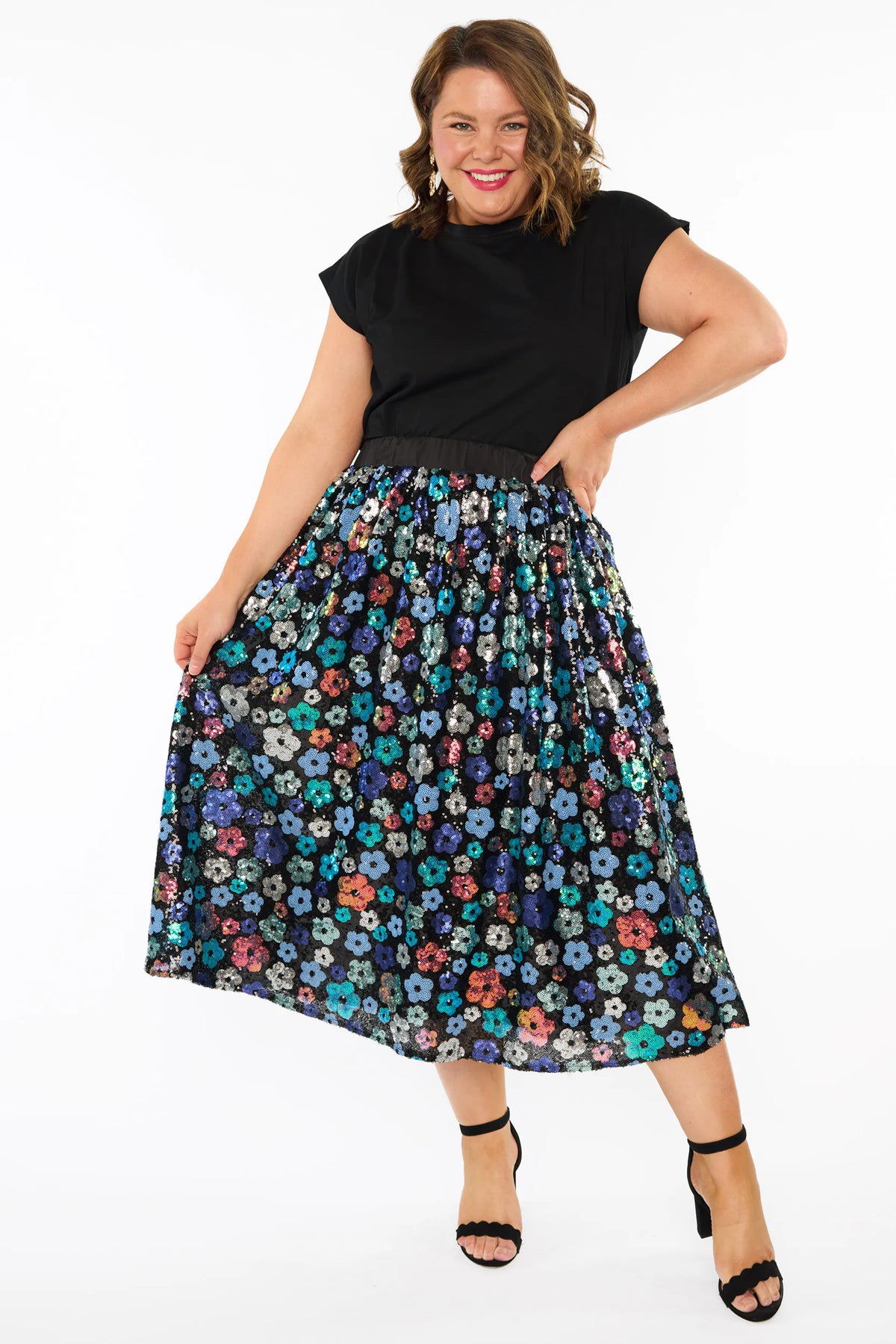 A woman modelling a short-sleeve black top, with a sequin knee length skirt featuring a mix of blue tonal coloured flowers on a black background. Paired with black heels, against a plain white studio background.