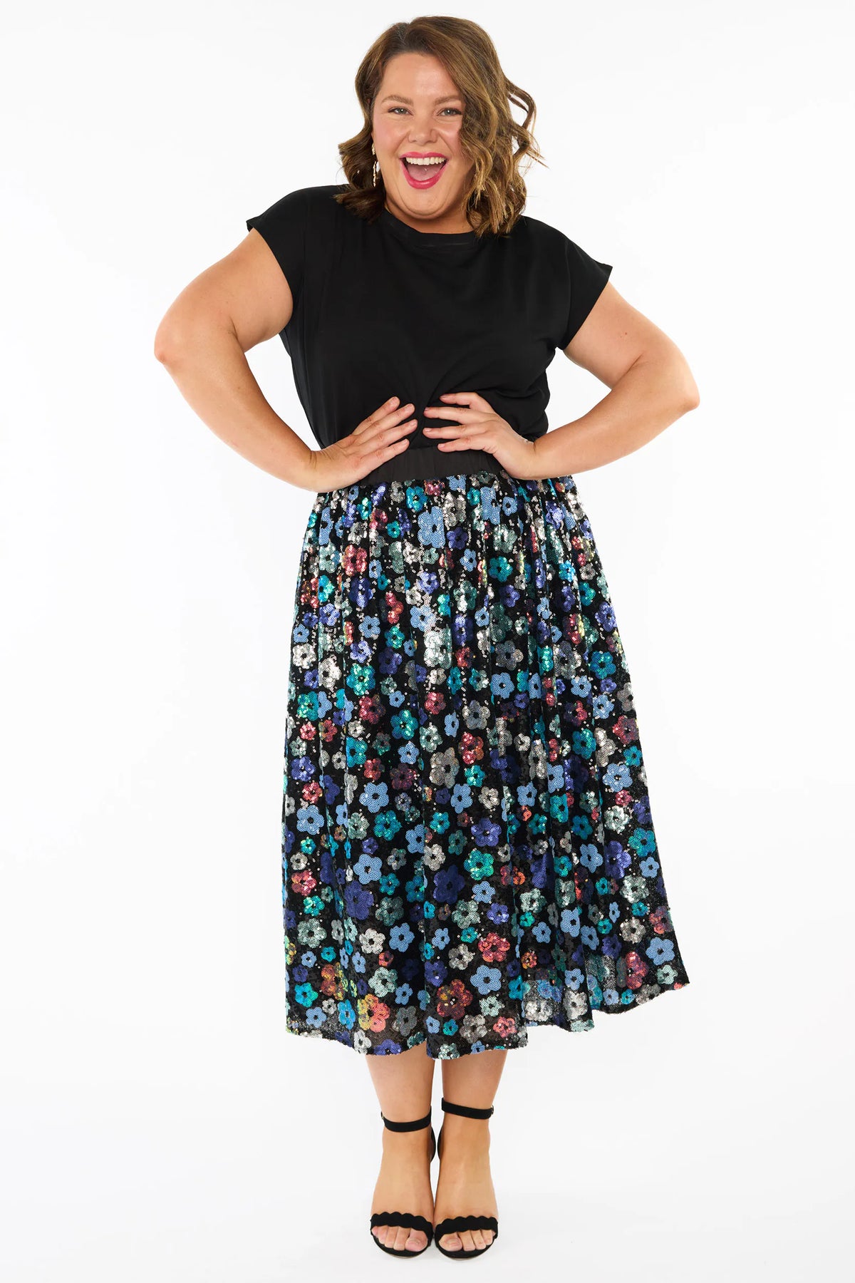 A woman modelling a short-sleeve black top, with a sequin knee length skirt featuring a mix of blue tonal coloured flowers on a black background. Paired with black heels, against a plain white studio background.
