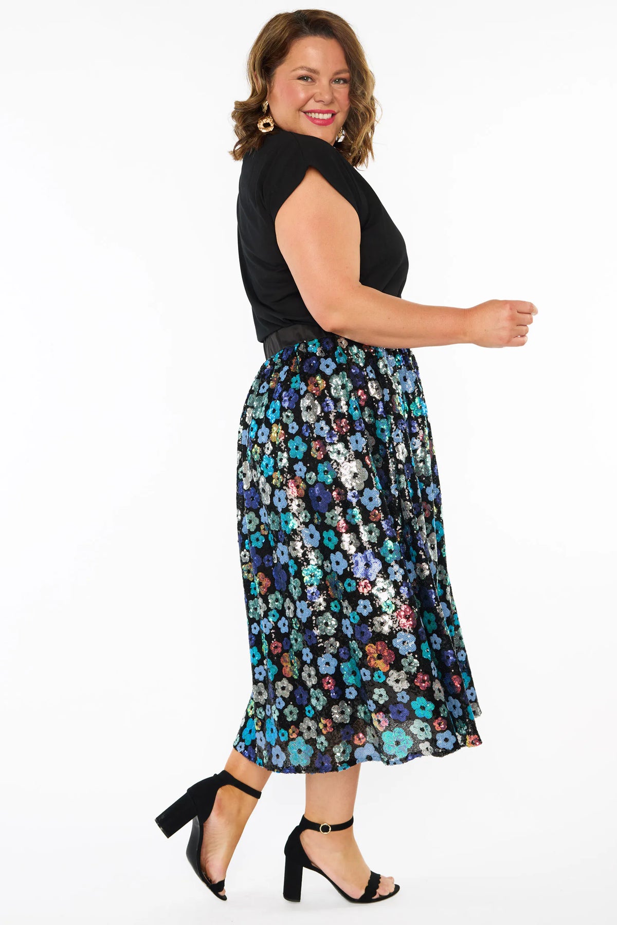 A woman modelling a short-sleeve black top, with a sequin knee length skirt featuring a mix of blue tonal coloured flowers on a black background. Paired with black heels, against a plain white studio background.