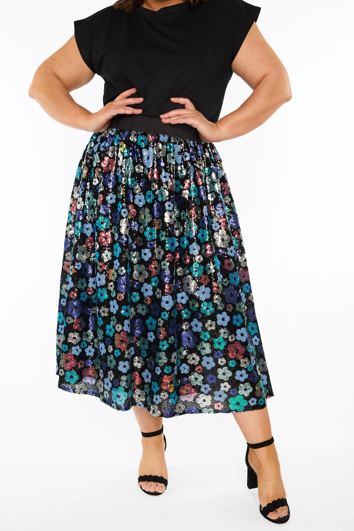 A woman modelling a short-sleeve black top, with a sequin knee length skirt featuring a mix of blue tonal coloured flowers on a black background. Paired with black heels, against a plain white studio background.