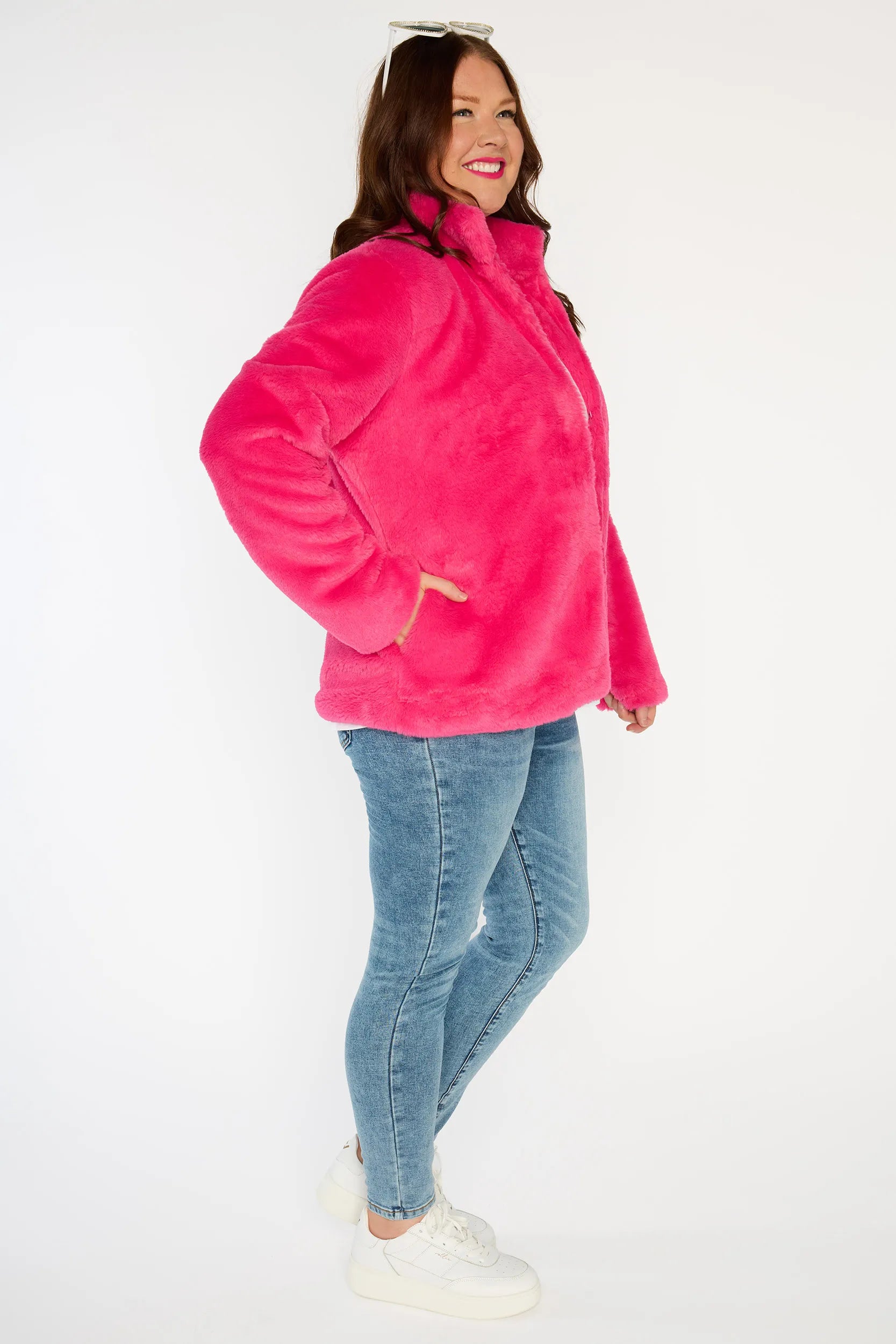 Person wearing a bright pink fleece jacket, white shirt, blue jeans, and white sneakers on a white background