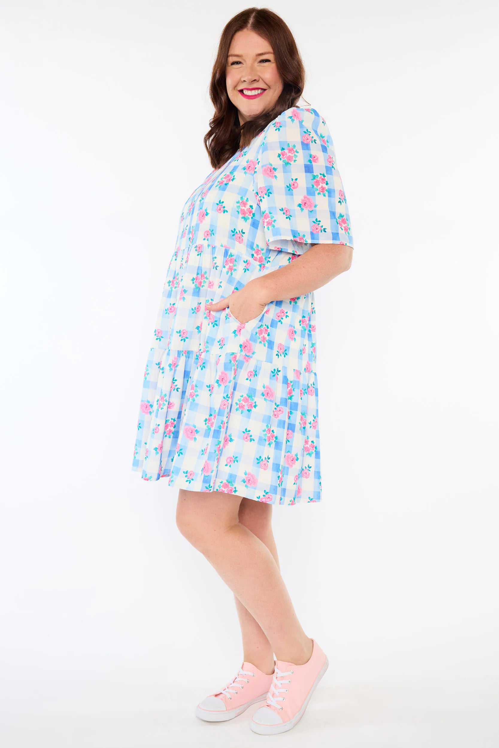 A woman modeling a short-sleeve, knee-length shift dress with a light blue, pink, and white floral print, paired with pink sneakers, against a plain white studio background.