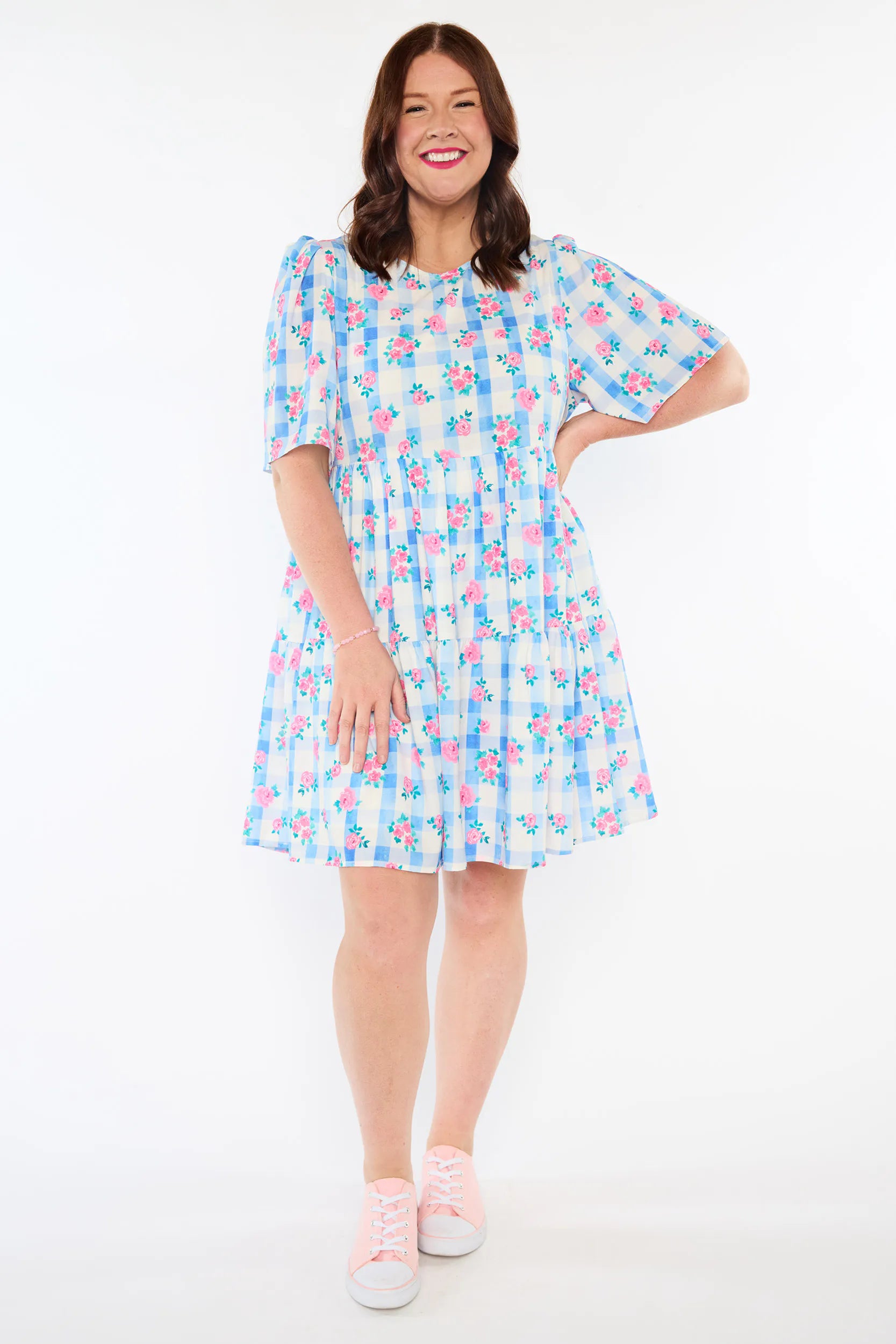 A woman modeling a short-sleeve tiered dress with a blue, pink, and white floral print, paired with light pink sneakers, against a plain white studio background.