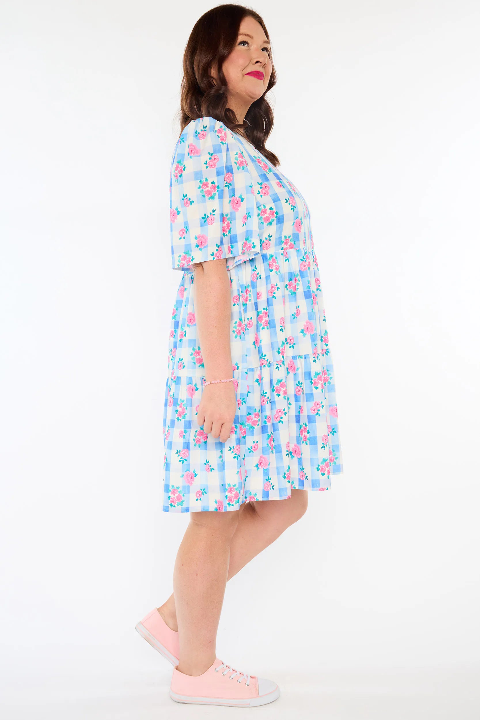 A woman modeling a light blue and white gingham dress with a pink floral pattern, paired with light pink sneakers, against a plain white studio background.