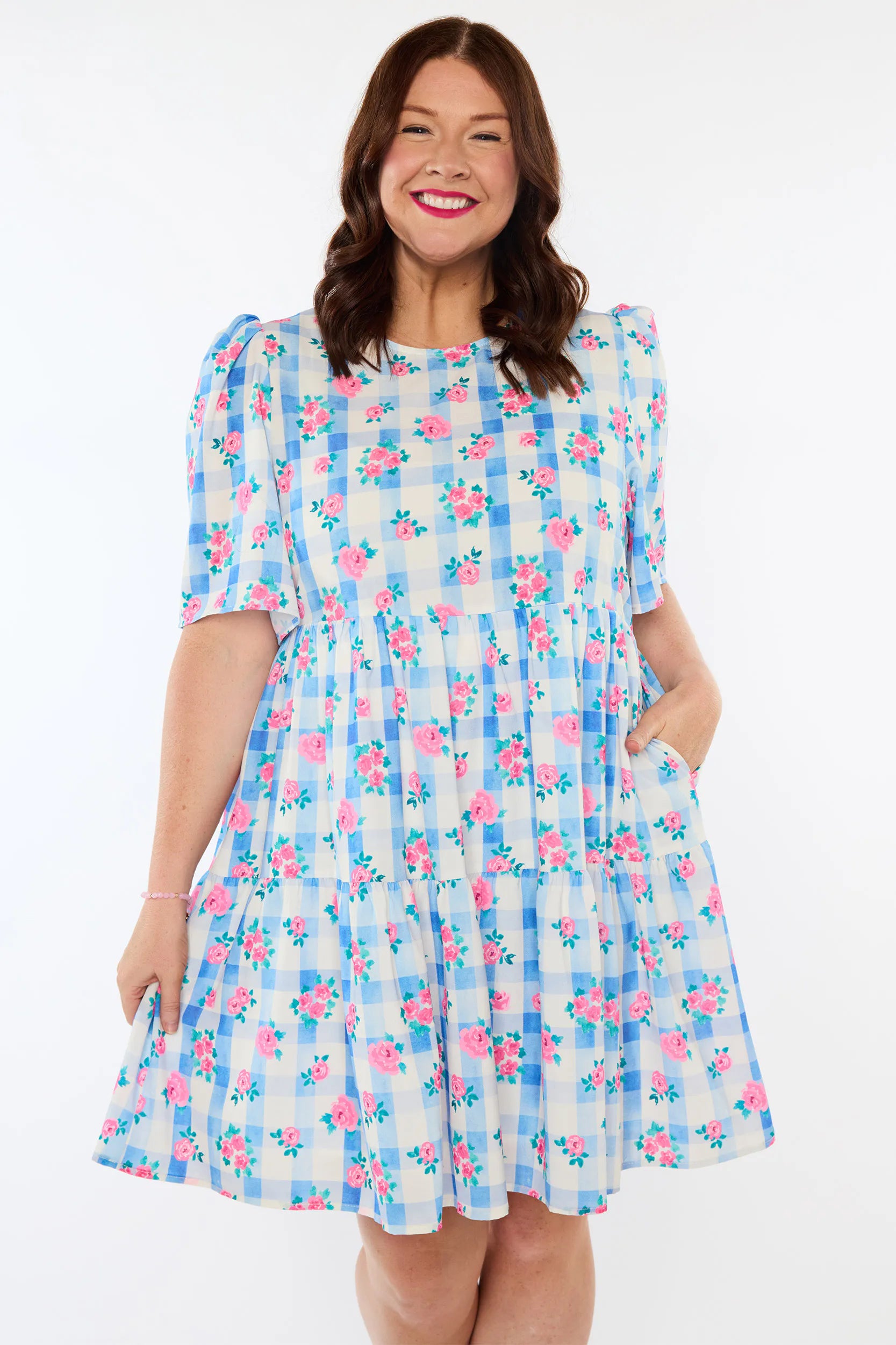 A woman modeling a short-sleeve tiered dress with a light blue and white gingham pattern and pink floral print, set against a plain white background.