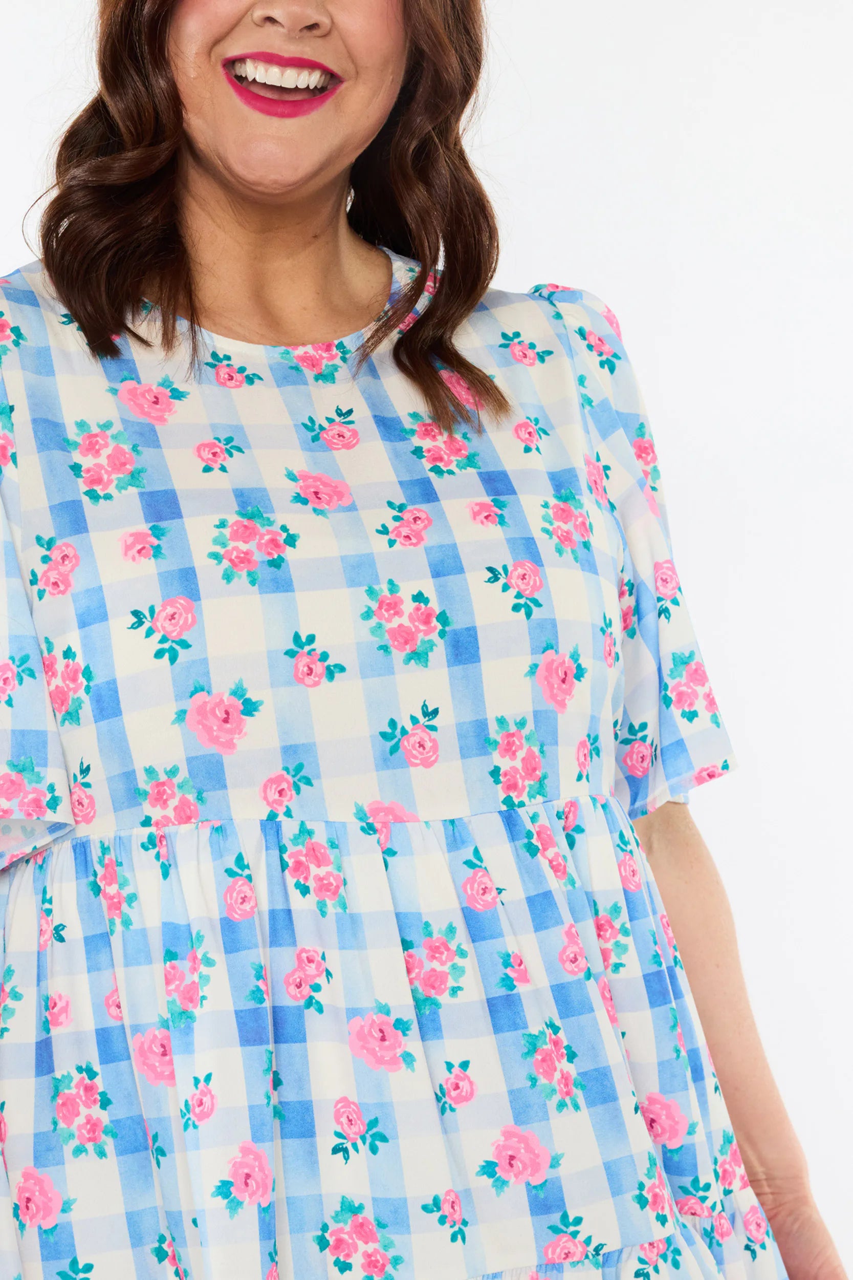 A close-up of a woman wearing a short-sleeve dress with a blue and white gingham check pattern overlaid with pink roses and green leaves, against a plain white background.