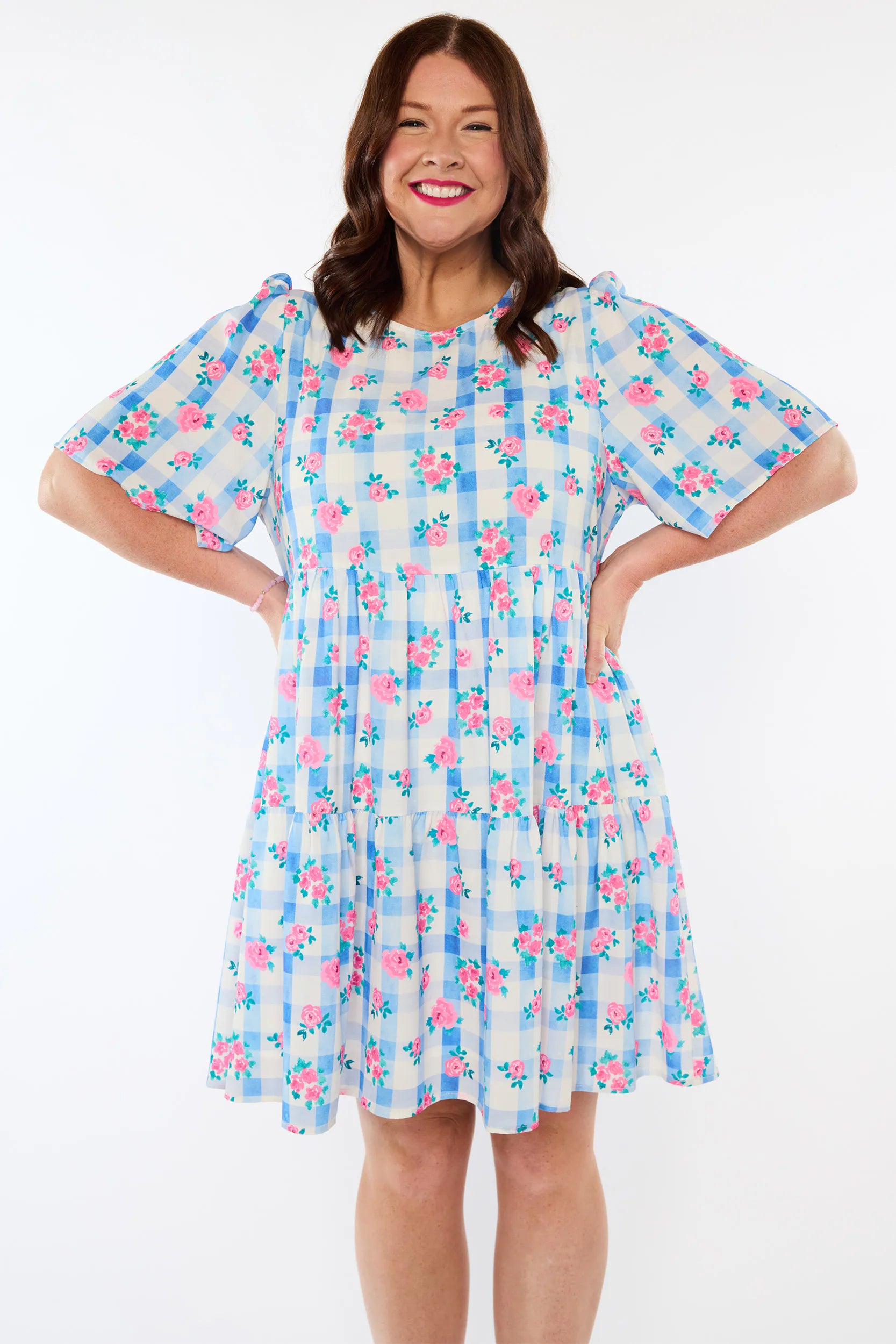 A woman modeling a short-sleeve tiered dress with a blue and white gingham pattern and pink floral print, set against a plain white background.