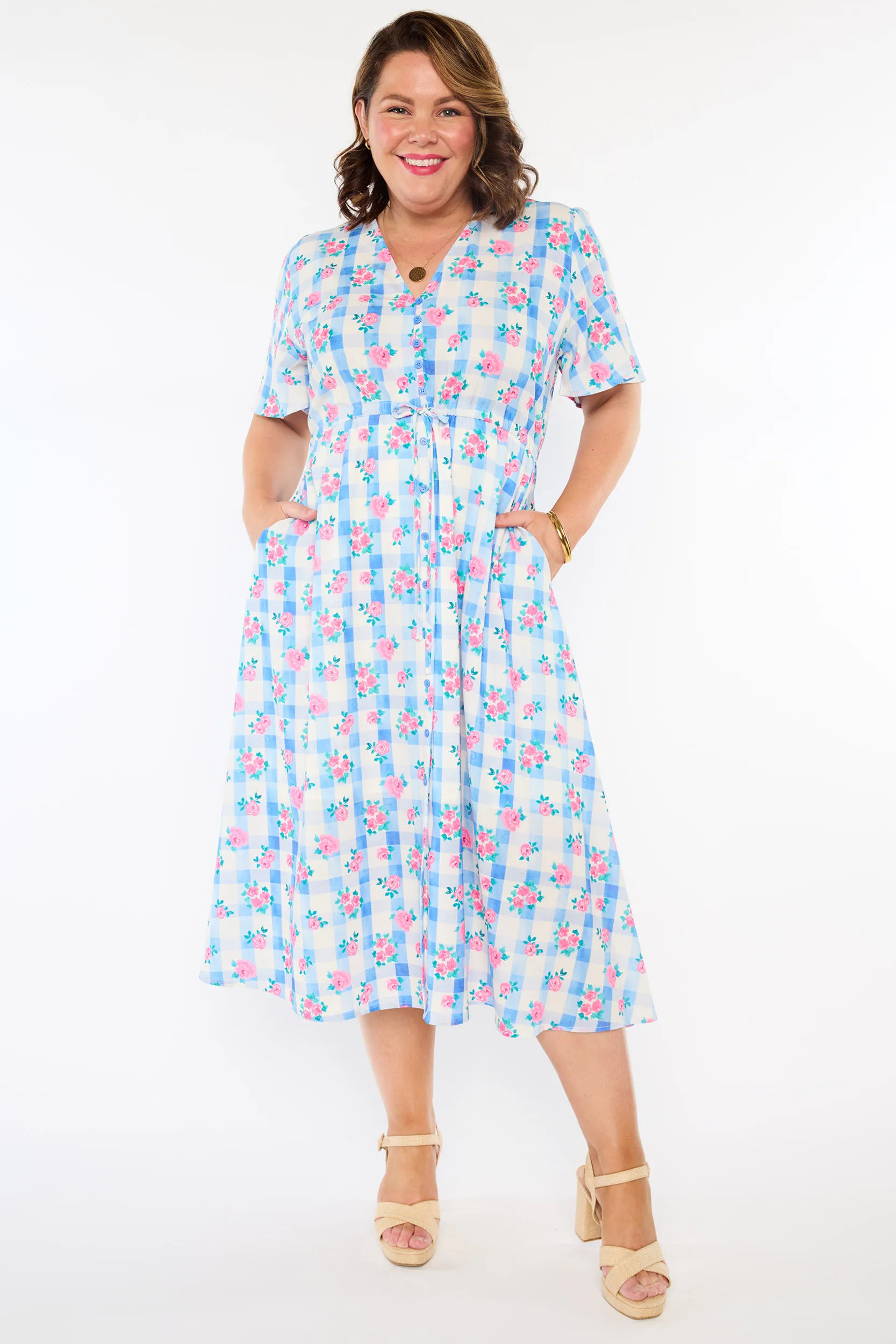 A woman modelling a short-sleeve, Midi-dress dress, with a button-up V-neckline & adjustable tie waist. A light blue and white gingham background covered in pink little roses, paired with cream heels, against a plain white studio background.