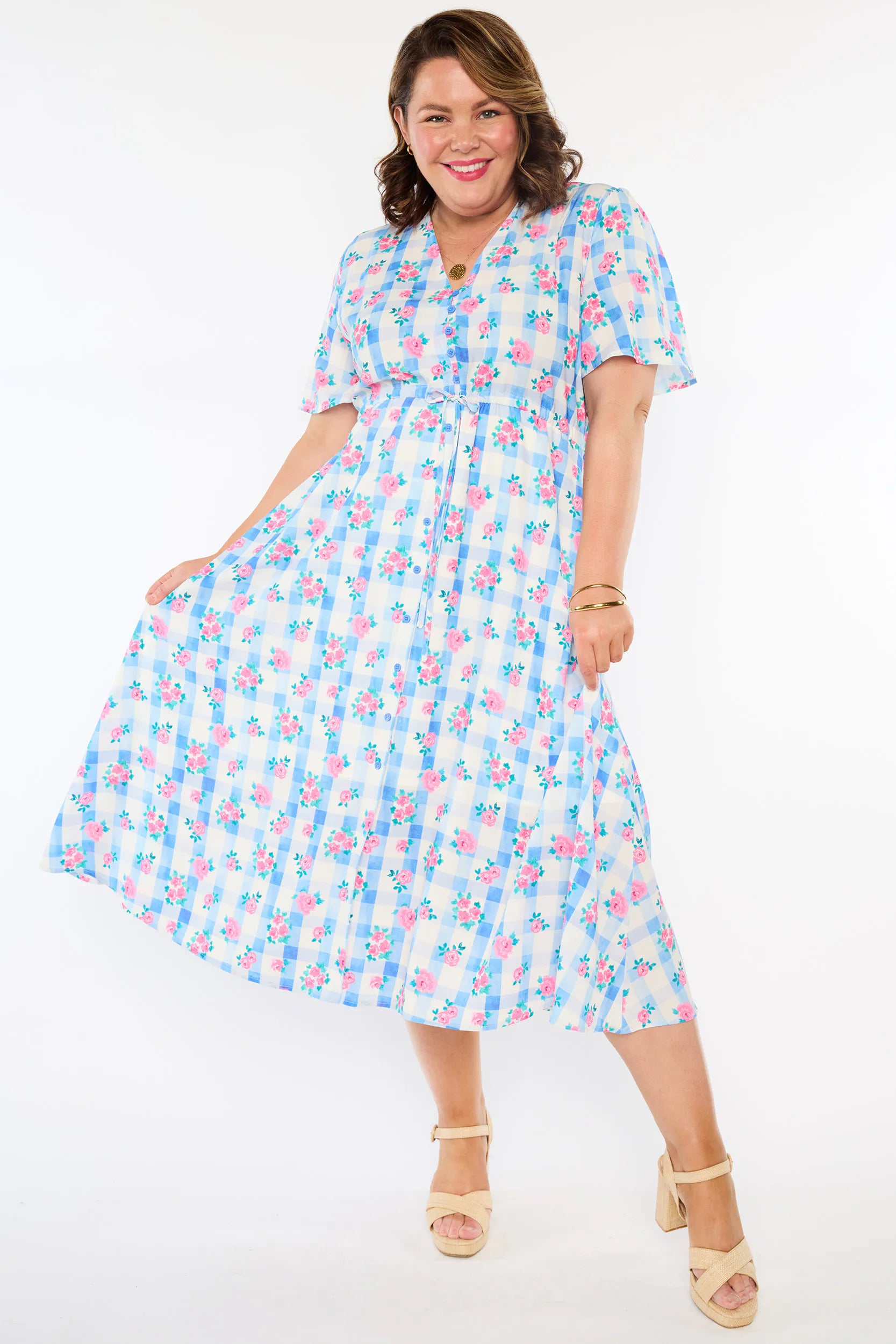 A woman modelling a short-sleeve, Midi-dress dress, with a button-up V-neckline & adjustable tie waist. A light blue and white gingham background covered in pink little roses, paired with cream heels, against a plain white studio background.