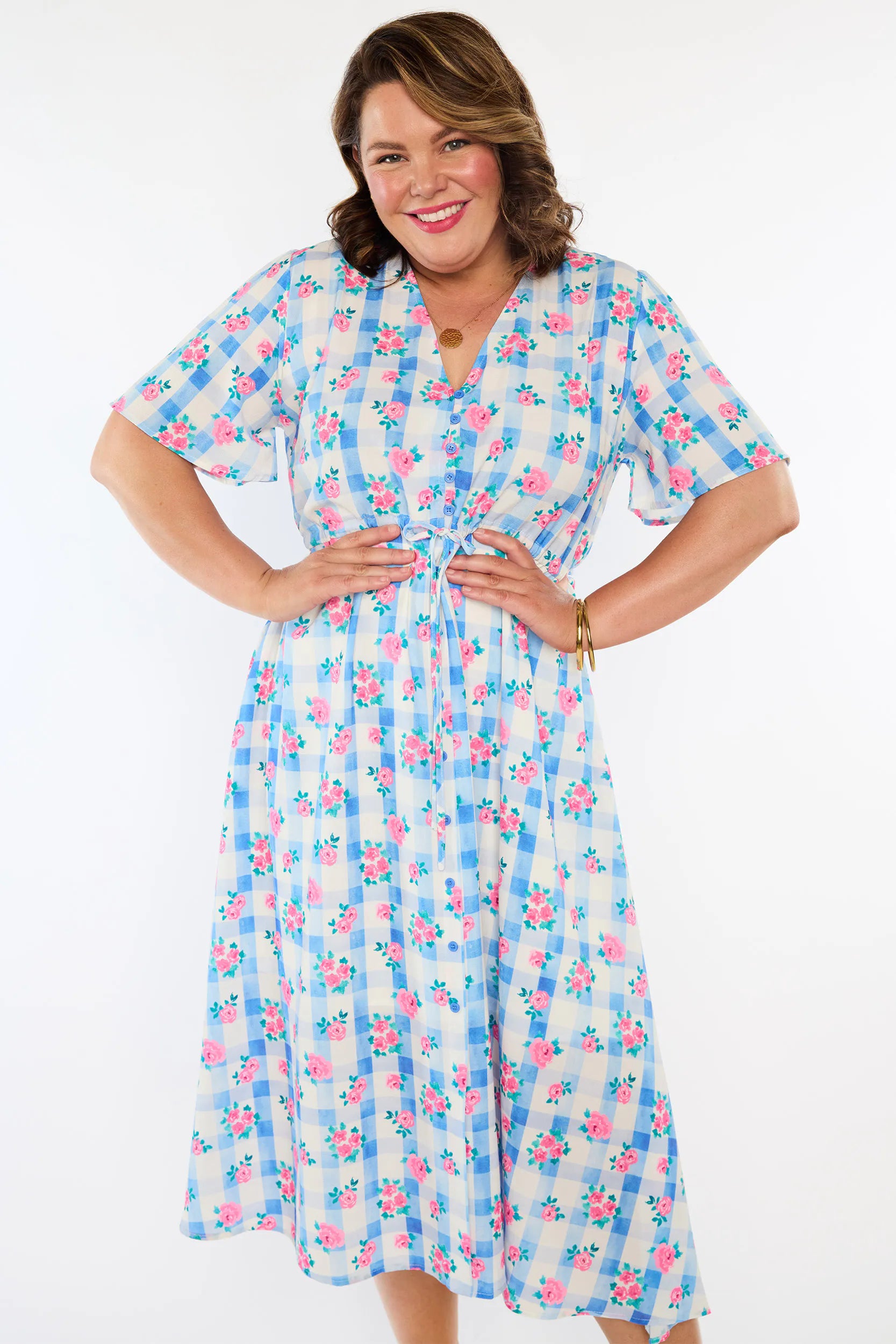 A woman modelling a short-sleeve, Midi-dress dress, with a button-up V-neckline & adjustable tie waist. A light blue and white gingham background covered in pink little roses, paired with cream heels, against a plain white studio background.