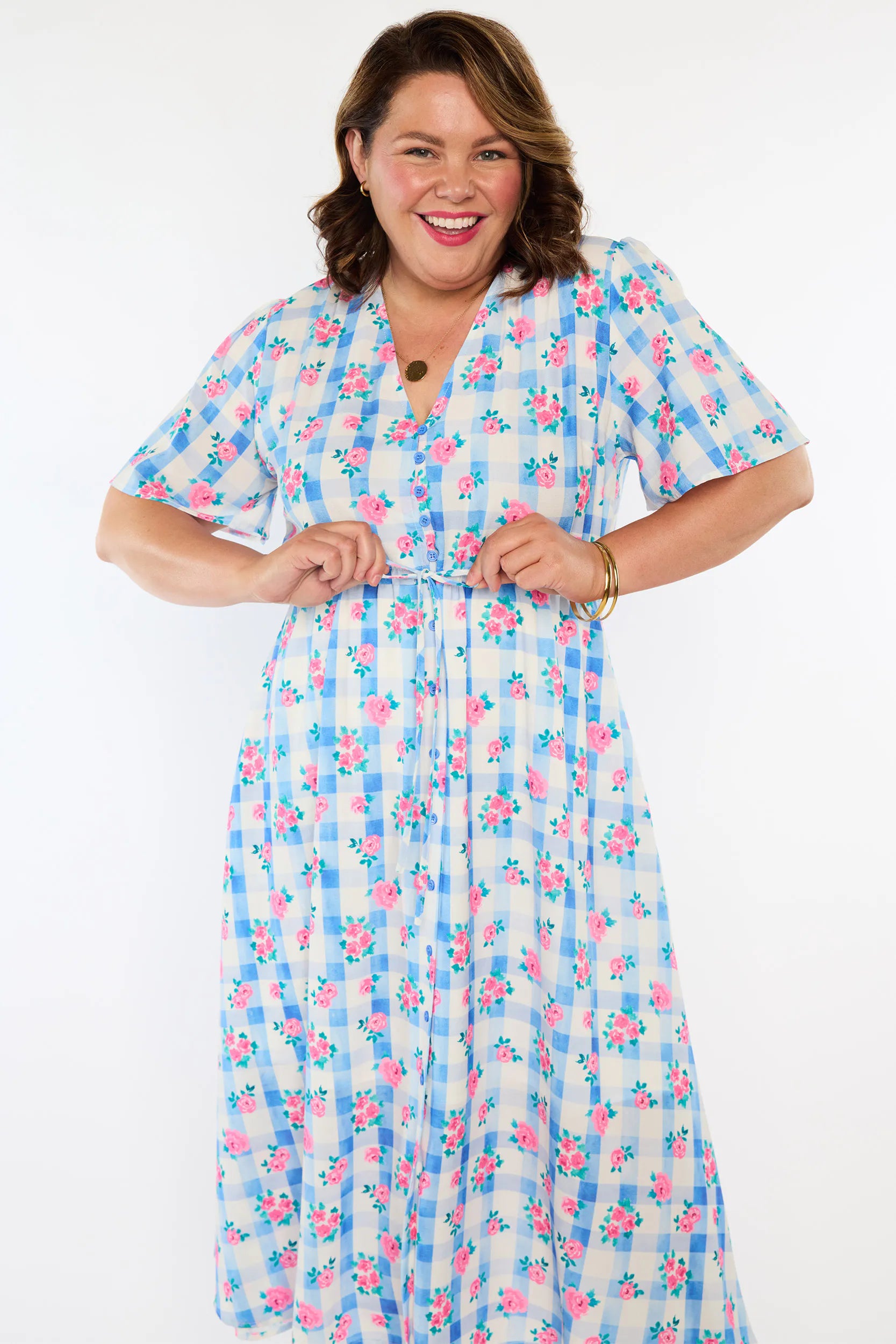A woman modelling a short-sleeve, Midi-dress dress, with a button-up V-neckline & adjustable tie waist. A light blue and white gingham background covered in pink little roses, paired with cream heels, against a plain white studio background.