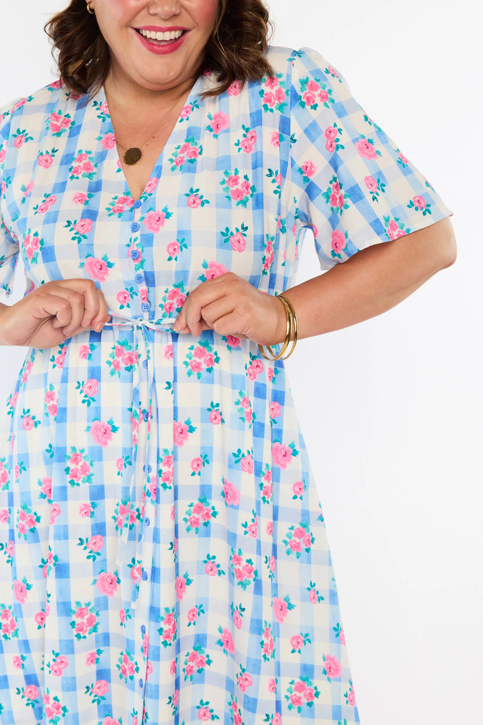A woman modelling a short-sleeve, Midi-dress dress, with a button-up V-neckline & adjustable tie waist. A light blue and white gingham background covered in pink little roses, paired with cream heels, against a plain white studio background.