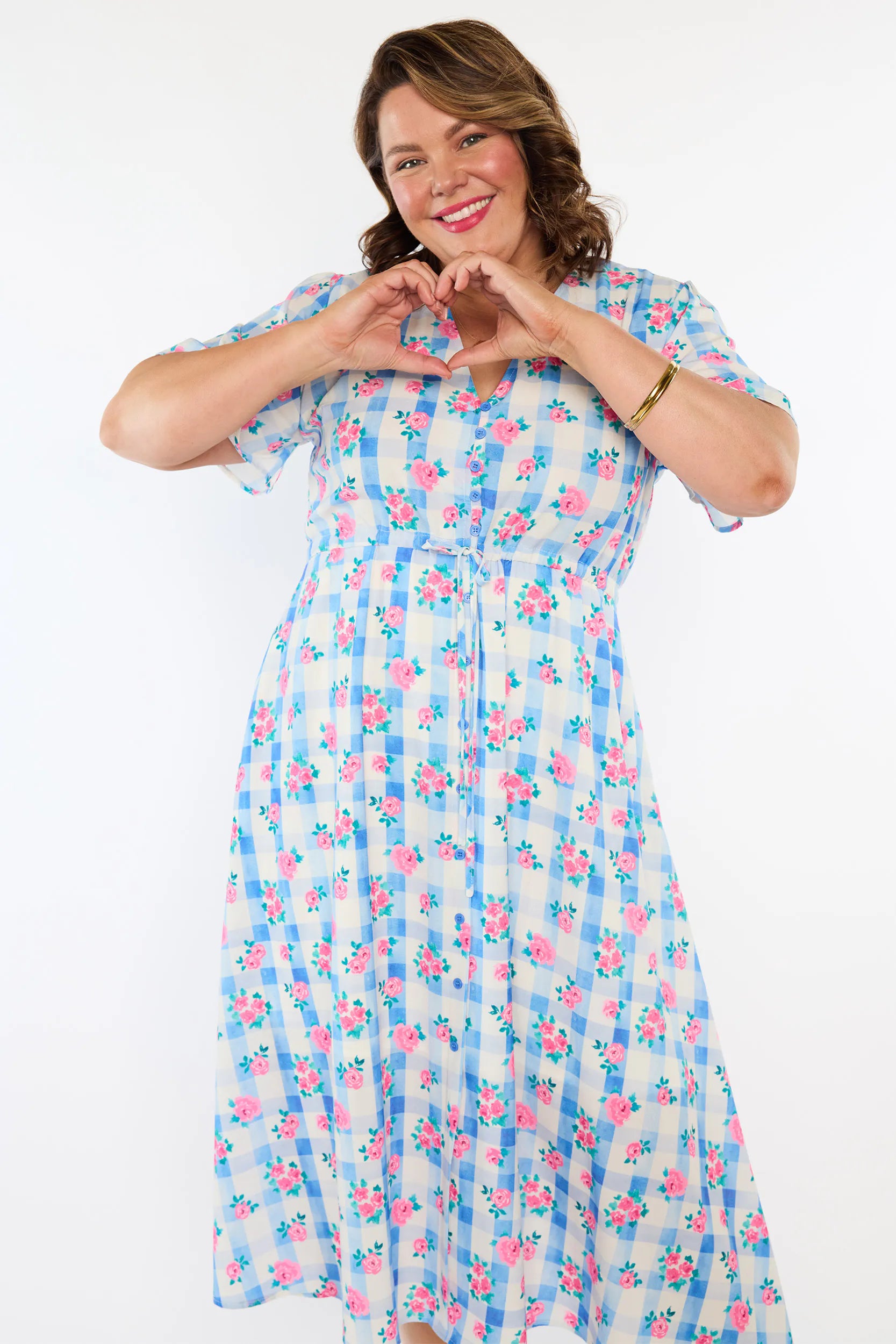 A woman modelling a short-sleeve, Midi-dress dress, with a button-up V-neckline & adjustable tie waist. A light blue and white gingham background covered in pink little roses, paired with cream heels, against a plain white studio background.