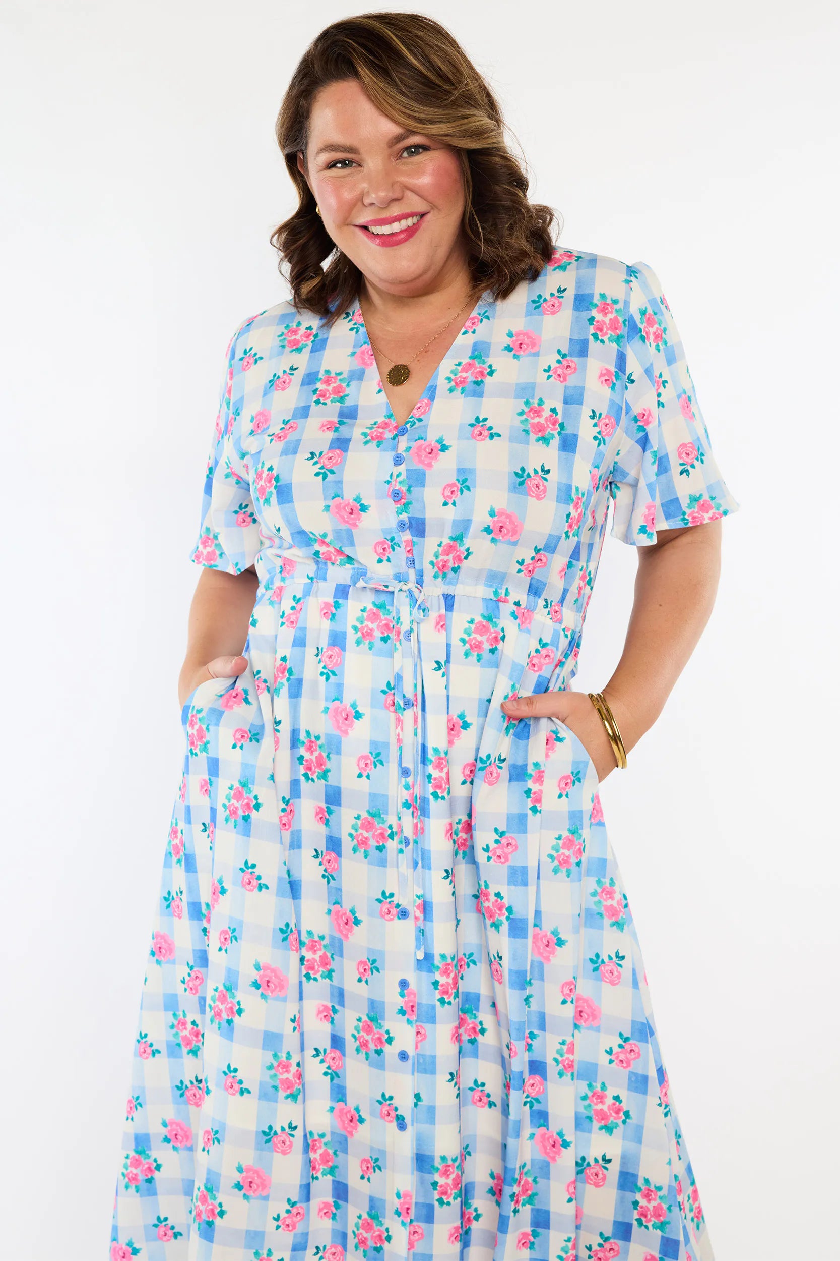 A woman modeling a short-sleeve midi dress with a blue and white gingham pattern and pink floral prints, featuring a V-neckline and side pockets.