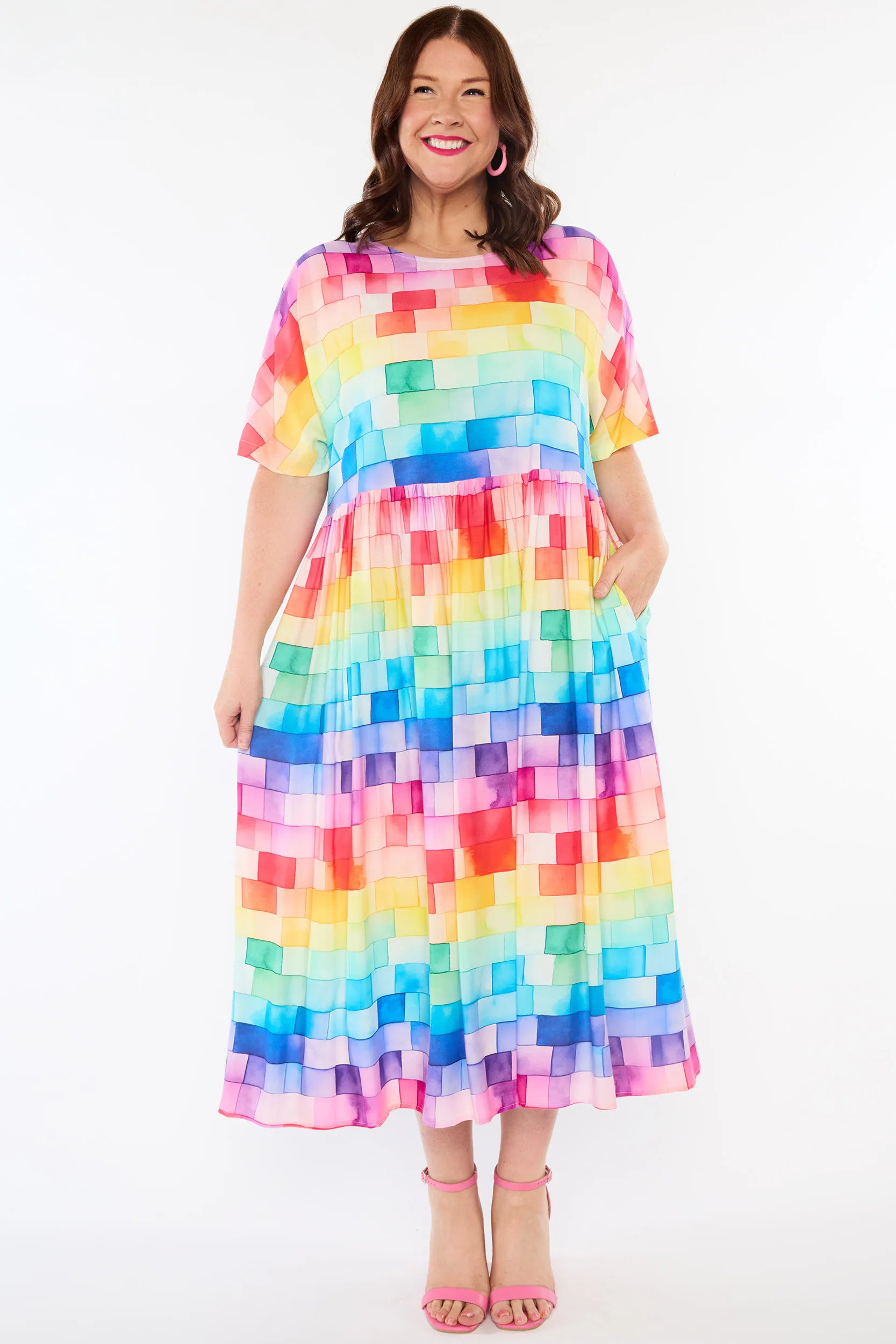 A woman modelling a short-sleeve midi dress with a colourful pixelated block pattern in shades of pink, orange, yellow, green, blue, and purple, paired with pink high-heeled sandals against a white studio background.