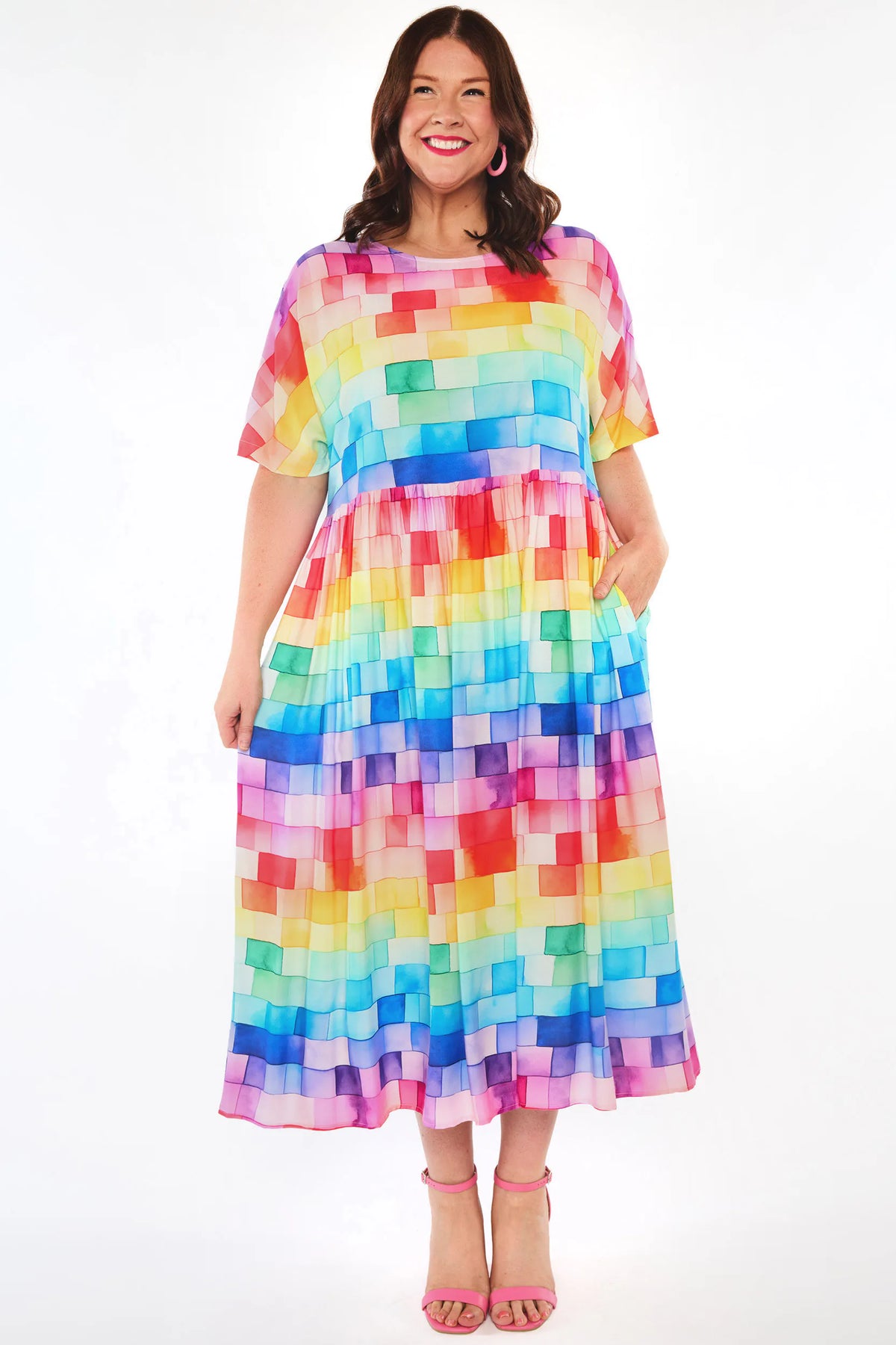 A woman modelling a short-sleeve midi dress with a colourful pixelated block pattern in shades of pink, yellow, green, blue, and purple, paired with pink high-heeled sandals against a white studio background.