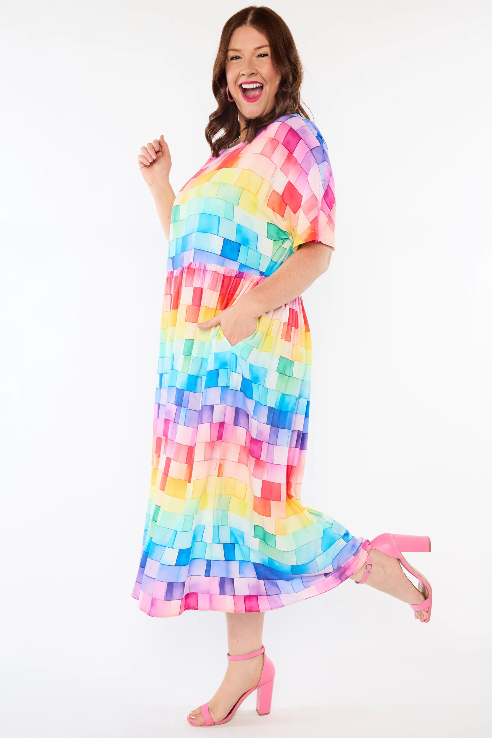 A woman modelling a short-sleeve midi dress with a colourful rainbow pixelated block pattern, paired with pink high-heeled sandals, against a plain white studio background.