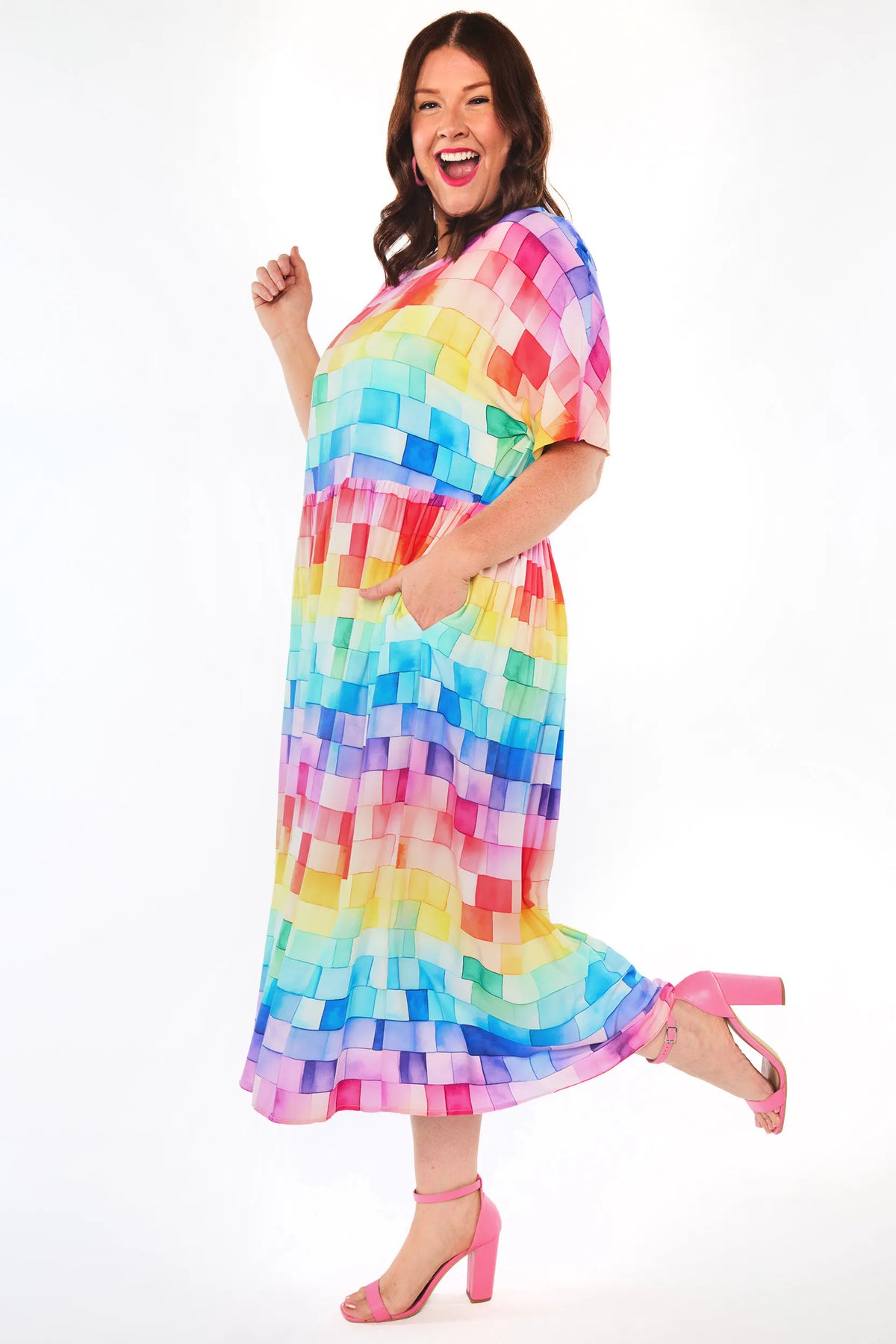 A woman modelling a short-sleeve midi dress with a colourful pixelated block pattern in shades of pink, blue, yellow, green, and orange, paired with pink high-heeled sandals against a white studio background.