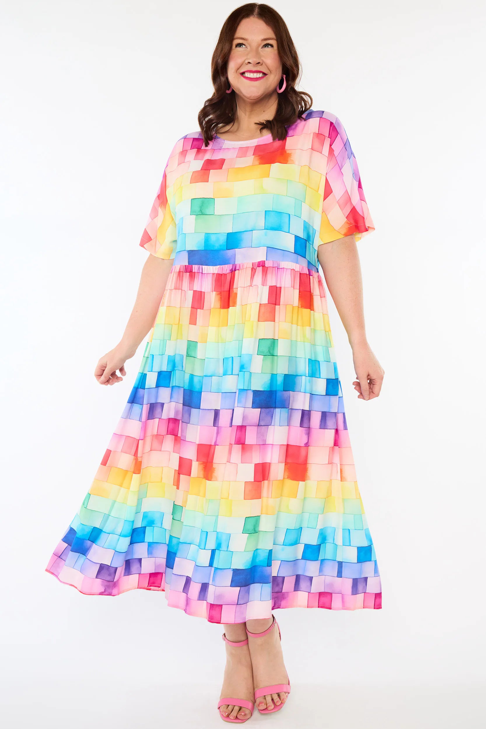 A woman modelling a short-sleeve midi dress with a colourful rainbow block pattern, standing against a plain white studio background.