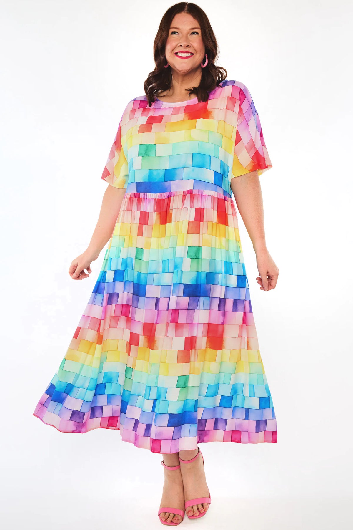 A woman modelling a short-sleeve midi dress with a colourful rainbow block pattern, standing against a plain white studio background.