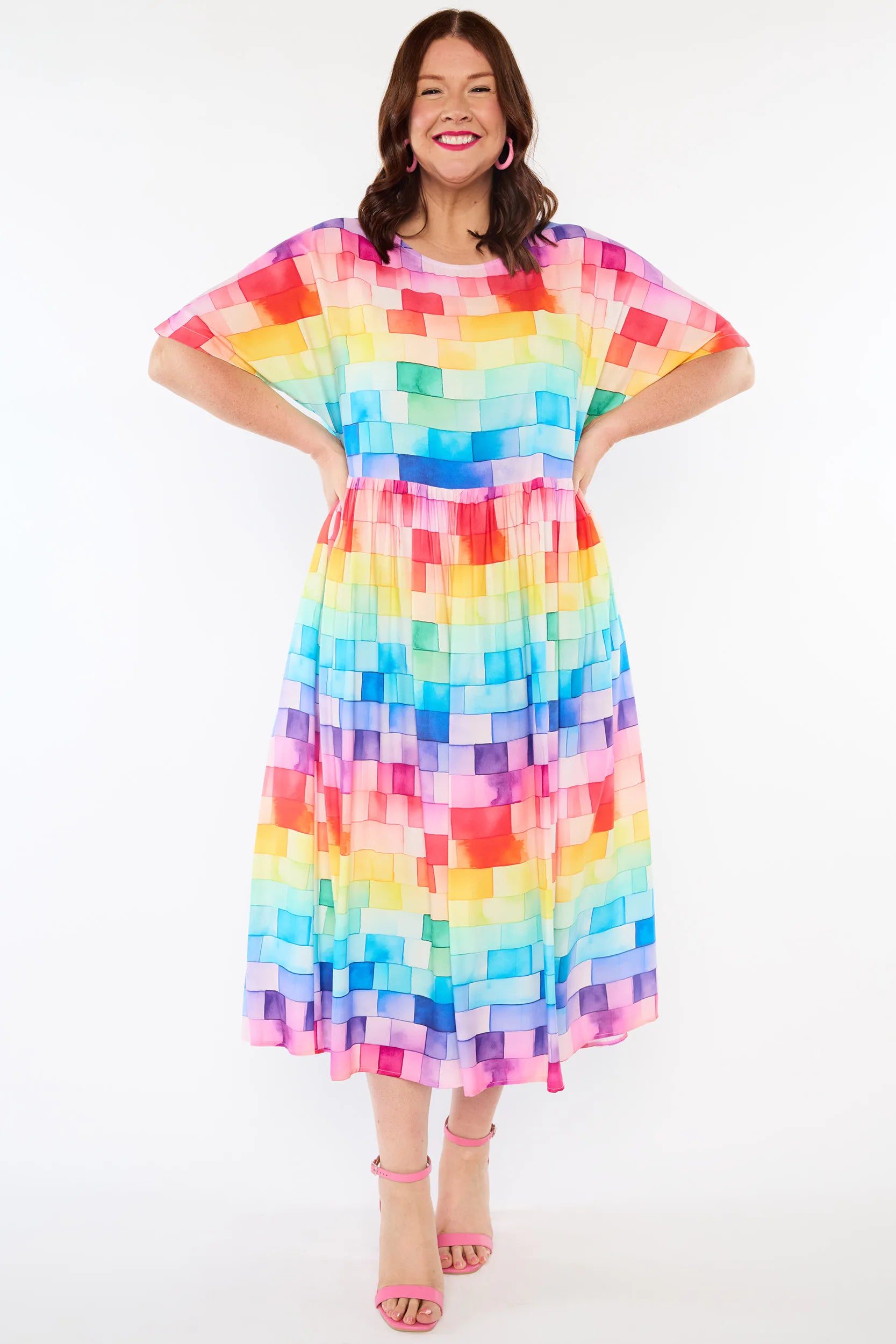 A woman modelling a short-sleeve midi dress with a colourful rainbow block pattern, standing against a plain white studio background.