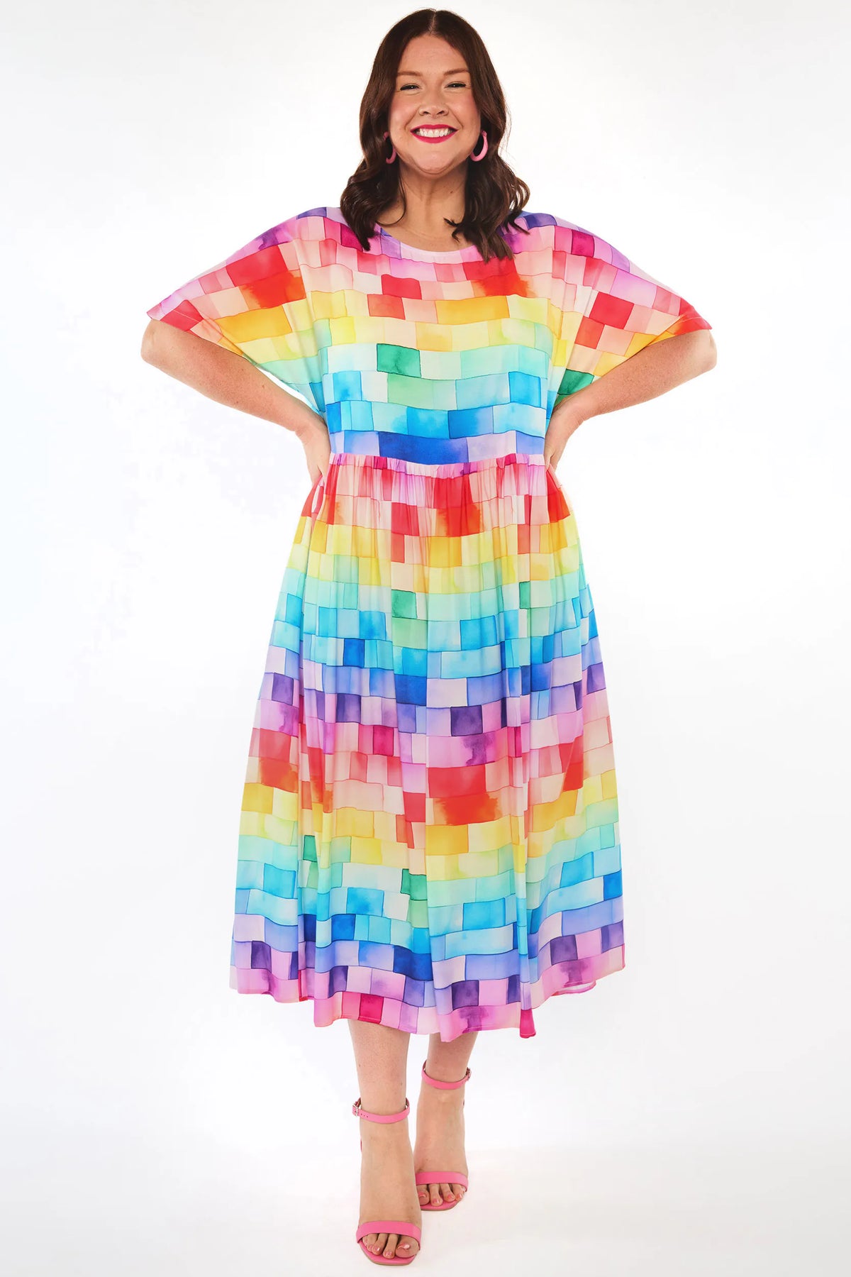 A woman modelling a short-sleeve midi dress with a colourful pixelated block pattern in shades of pink, red, orange, yellow, green, blue, and purple, paired with pink high-heeled sandals against a white studio background.